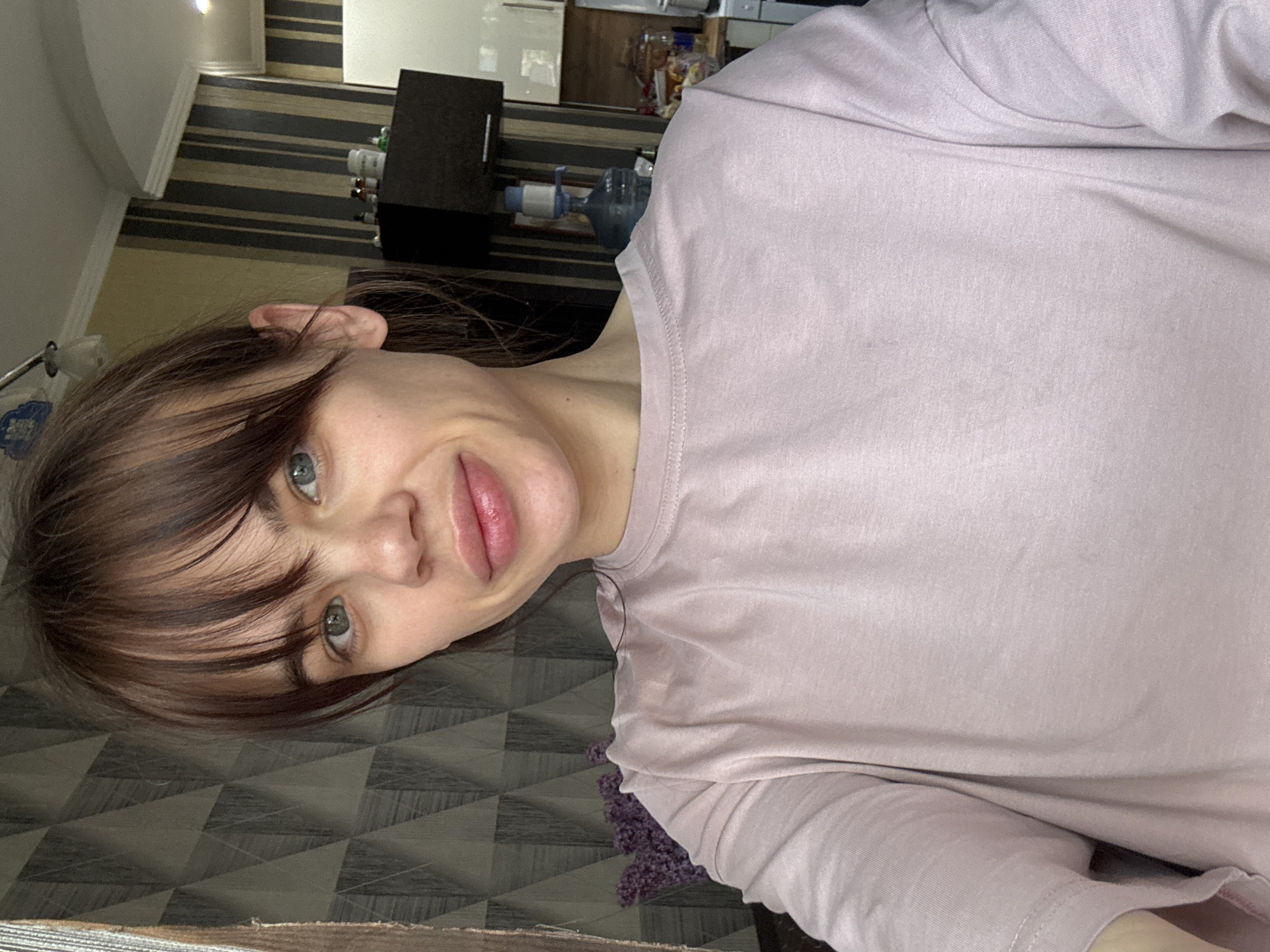 A woman with brown hair and bangs, wearing a pink t-shirt, is posing for a selfie indoors. The background includes a wall with striped wallpaper and a cabinet. A water cooler and other items are visible in the background. It is not possible to determine the specific location in the city of Rostov-on-Don, Russia from the image.