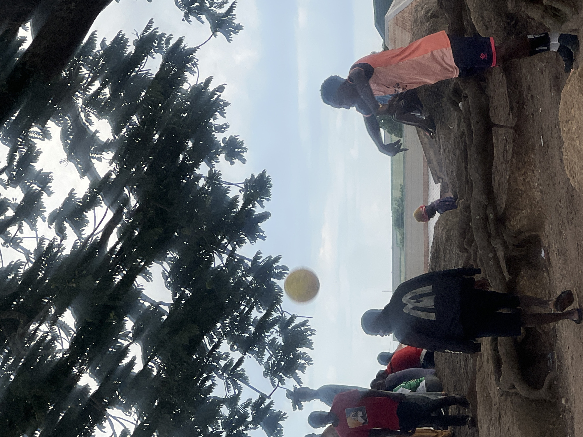 The image captures a group of people in what appears to be an outdoor gathering in Jos, Nigeria. A large tree with dark green foliage dominates the upper portion of the frame, with sunlight filtering through its branches, creating dappled lighting on the scene below.
In the mid-ground, a yellow ball is suspended in the air, suggesting a game or activity is in progress. Several individuals are visible. To the left, a man in a red t-shirt stands, facing away from the camera. Several other people are clustered behind him, some seated, others standing.
In the foreground and mid-ground to the right, a young man in an orange and pink tank top and dark shorts is seated on a rock formation, looking to his left. His posture suggests a moment of rest or observation. Behind him, another individual is partially visible.
Further in the background, a person wearing a black jacket with a prominent white McDonald's logo on the back is walking away from the camera. They are wearing dark shorts and flip-flops.
The ground appears to be rough and earthy, with exposed tree roots visible in the foreground, indicating a natural or semi-natural setting. The sky is overcast with some patches of blue, suggesting a cloudy day. In the far distance, there are hints of buildings or structures, possibly indicating the edge of a settlement or a more developed area within Jos. The overall atmosphere is casual and communal.