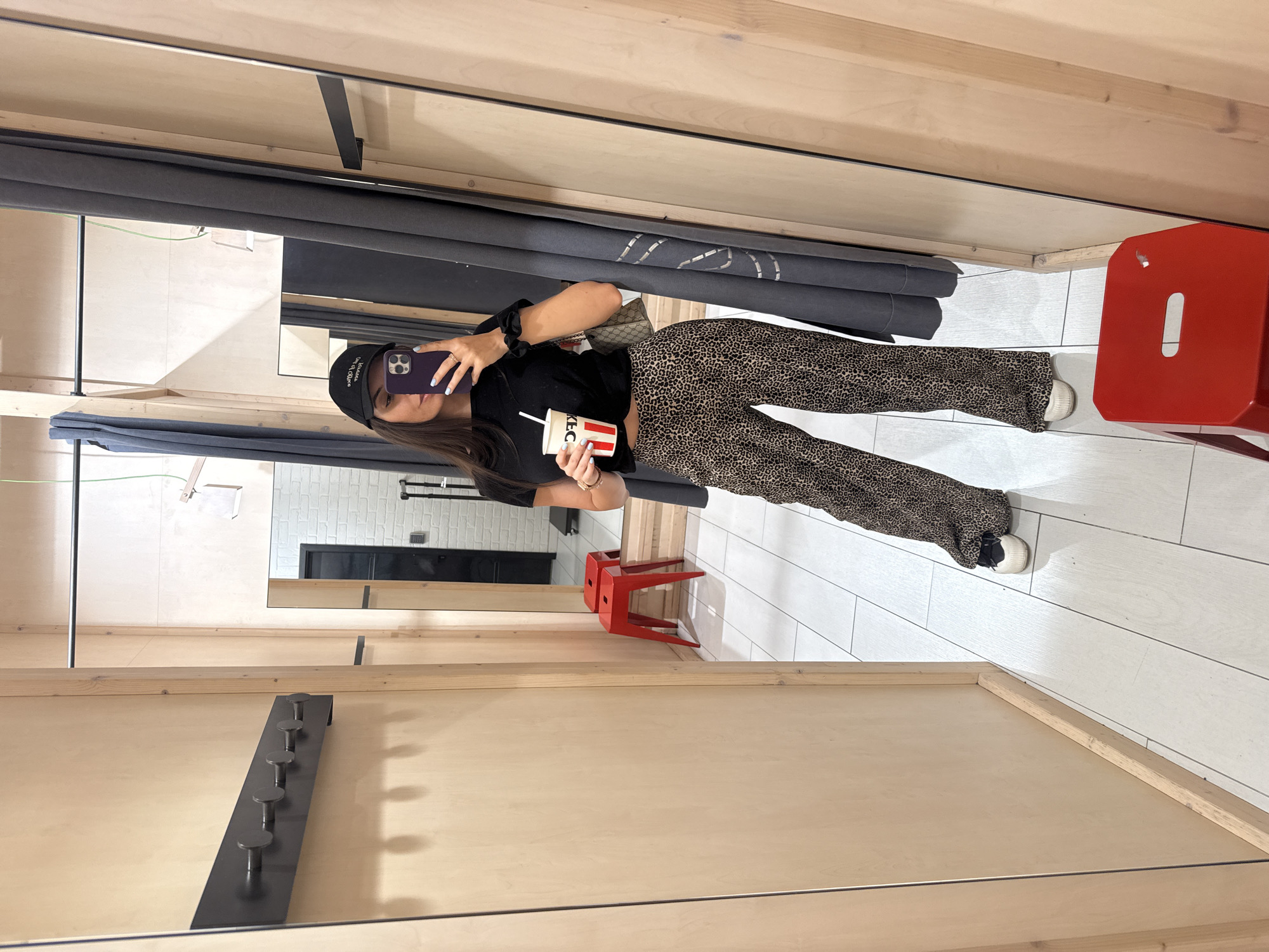 A person in a dressing room is taking a selfie in a mirror. The person is wearing a black cap, black shirt, and leopard print pants. They are holding a cup with "KFC" printed on it in their left hand and a purple phone in their right hand. A red stool is visible in the corner. The changing room has a wooden frame and a gray curtain. The reflection shows an additional room behind them with a white brick wall and a black cabinet. There is no definitive evidence to place this scene within the city of Plovdiv, Bulgaria.