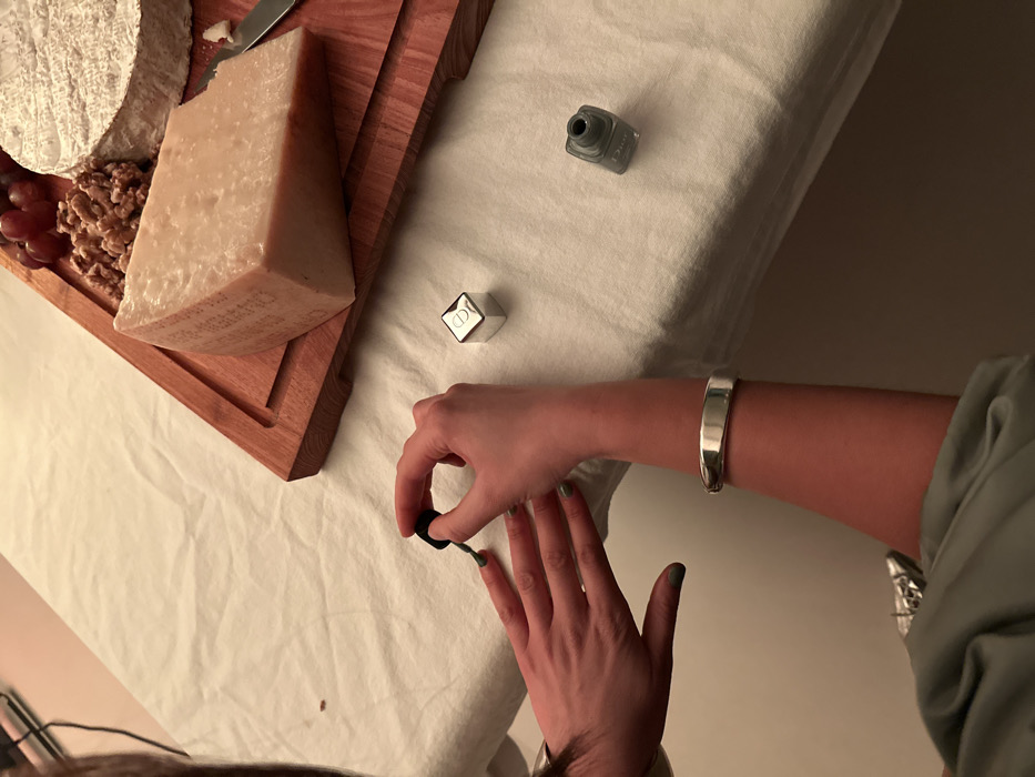 The image displays an overhead view of a person's hands engaged in applying nail polish. The individual's right hand holds a brush, painting the fingernails of the left hand with a green-grey polish. A polished silver bracelet is visible on the person's right wrist. The scene takes place indoors, with a white tablecloth covering a surface. On the table, to the right of the hands, rests a wooden cutting board laden with food items. These include a large wedge of Parmesan-style cheese, a round soft cheese, a cluster of red grapes, and several walnuts. A small silver knife is positioned on the board near the Parmesan. Additionally, a small, silver cubic object embossed with a "CD" logo (Dior) and an uncapped bottle of green-grey nail polish are present on the white tablecloth. The specific location within the city of London, United Kingdom, cannot be determined from the visual information provided in the image.