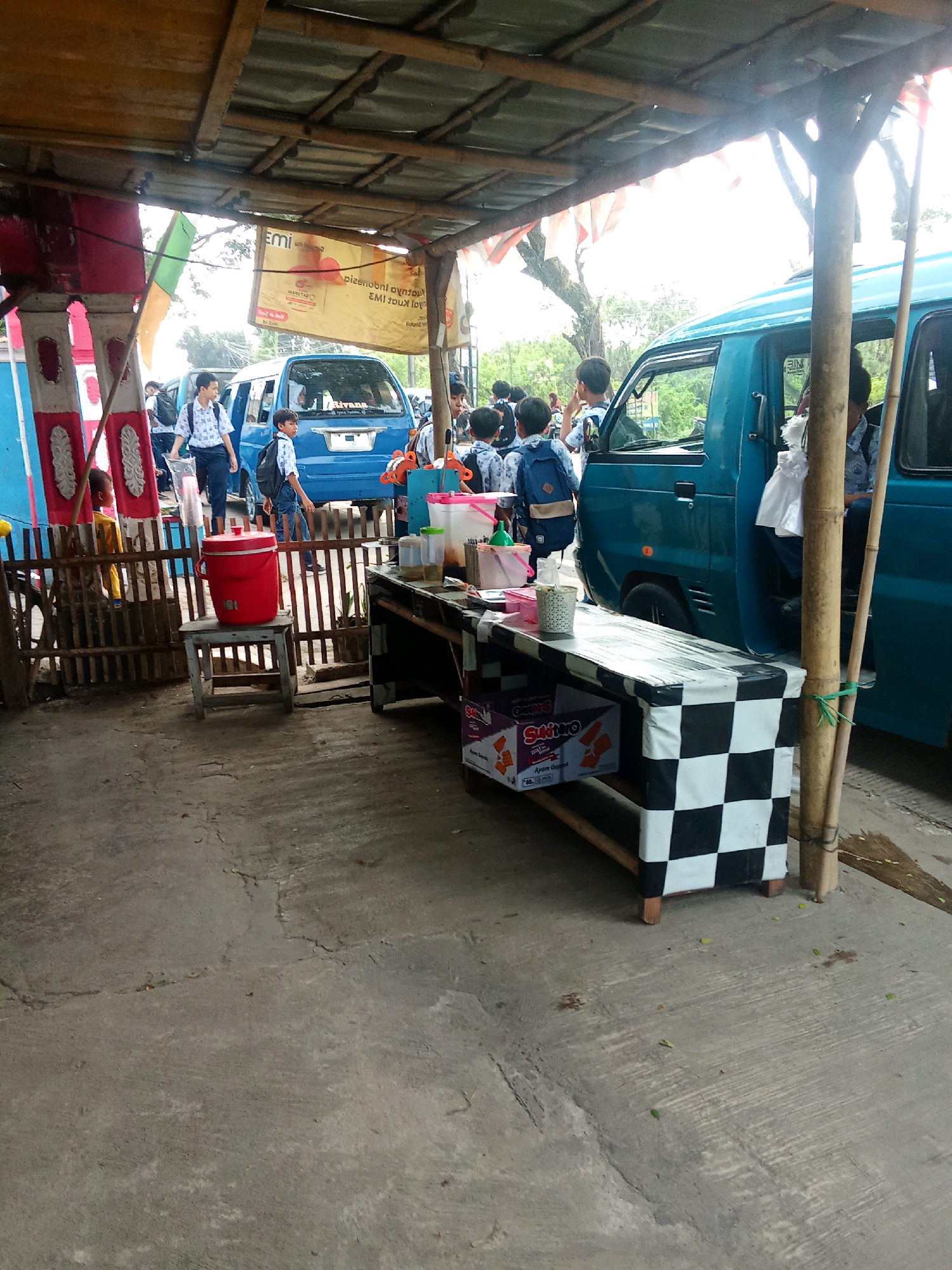 In Warunggunung, Indonesia, at what appears to be a roadside food stall or waiting area, children in school uniforms are gathered near two blue minivans. The scene is under a makeshift wooden and corrugated metal roof. A black and white checkered table displays drinks and snacks, with a red cooler on a small wooden stool in the foreground. The ground is concrete, with some debris. The overall impression is of a busy stop, likely near a school, where students are waiting for transportation.