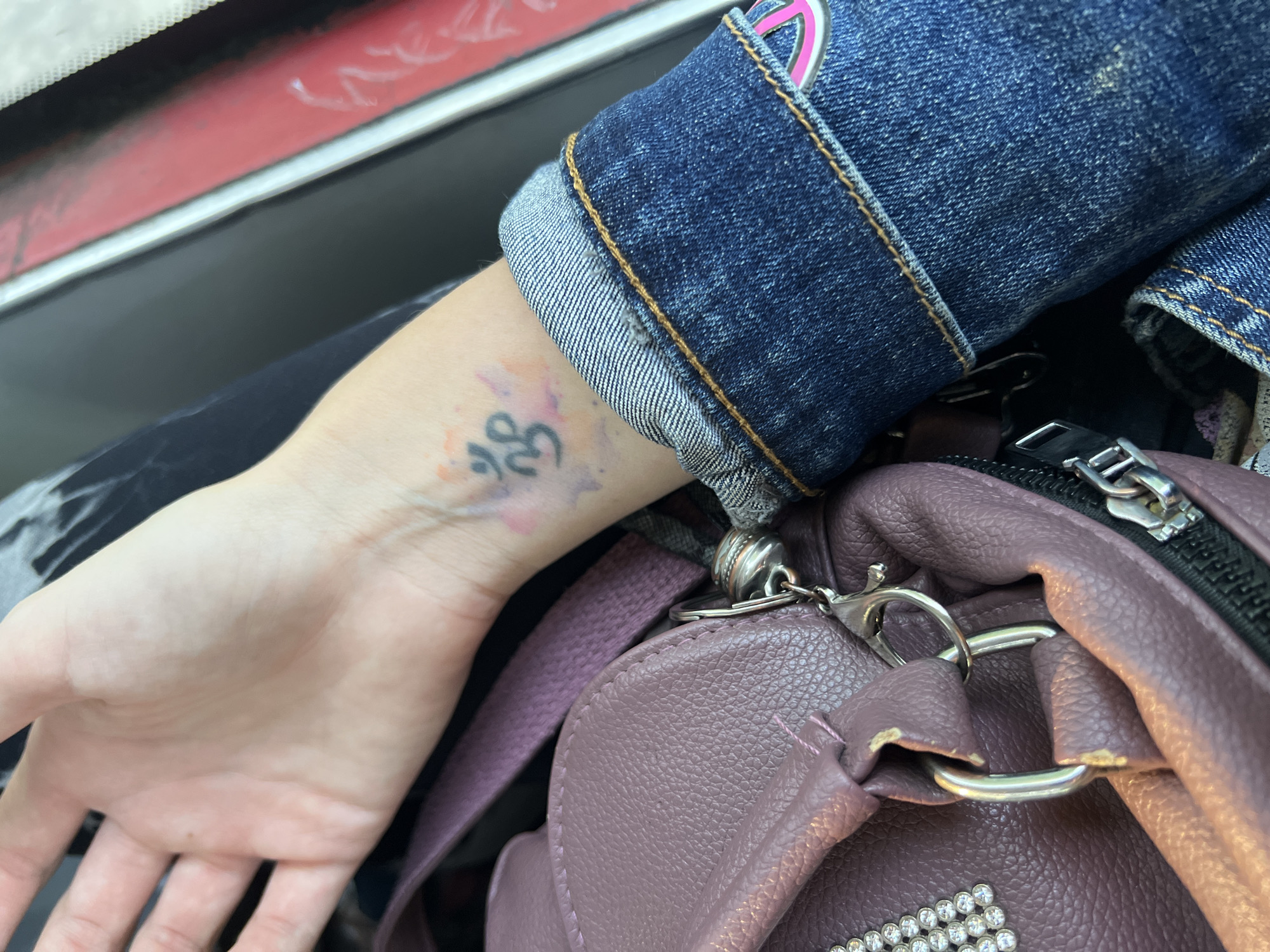 The image shows a close-up view of a person's left wrist, which has a tattoo of the Om symbol. The wrist is adorned with a small, dark Om symbol and some faded watercolor-like splotches of pink and orange. The person is wearing a blue denim jacket with visible stitching. Next to the wrist is a portion of a dusty rose-colored bag with metallic hardware and some shiny embellishments. The background is blurred, but suggests an indoor setting with elements of dark fabric and a hint of red. The location in the city of Лопаткино, Russia, is not discernible from the provided image.