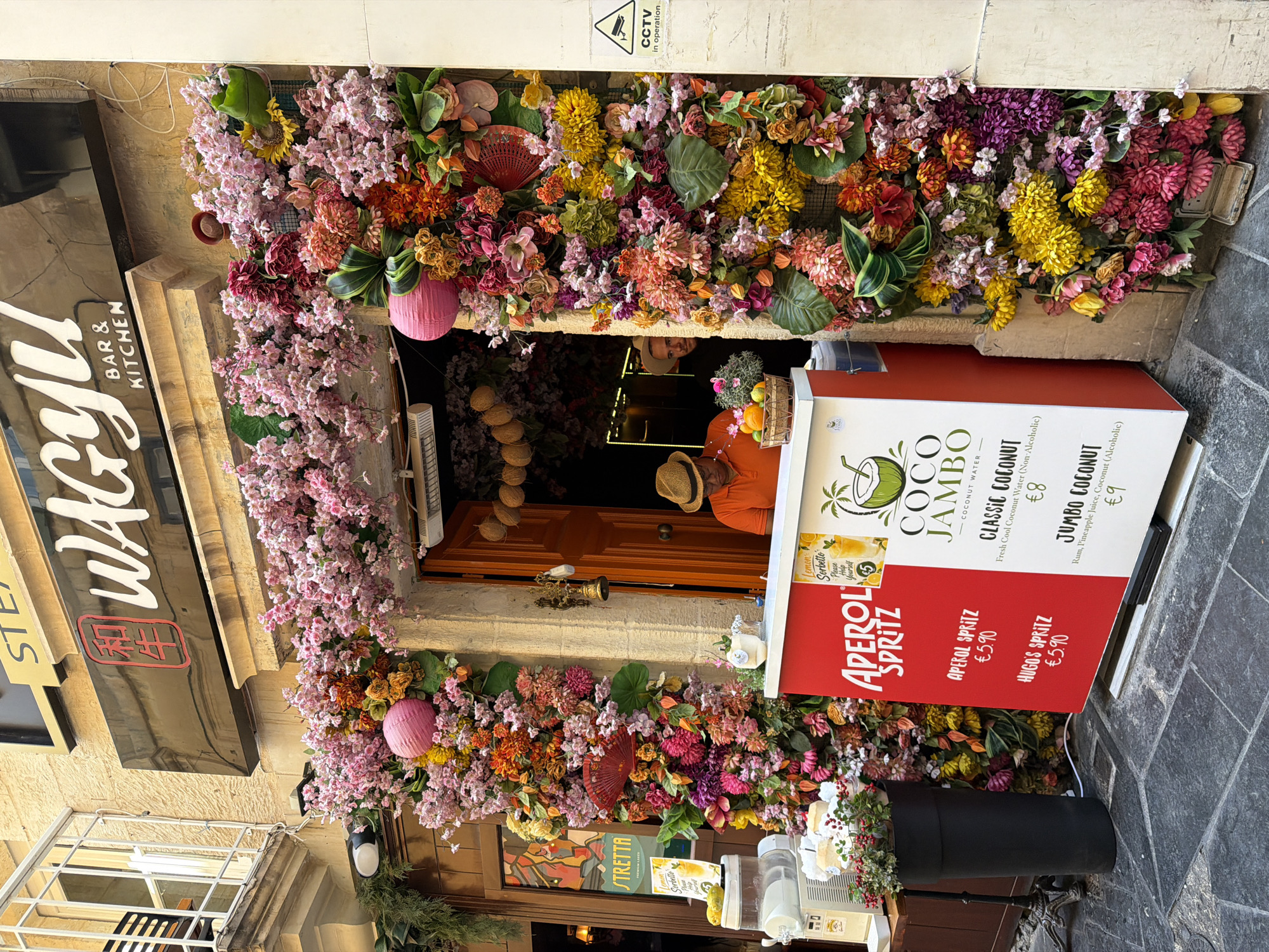 The image depicts an outdoor scene in Valletta, Malta, characterized by a vibrant floral arrangement adorning the entrance of a establishment. The flowers are a riot of colors, including pinks, oranges, yellows, and purples, creating a festive and inviting atmosphere.
A person wearing an orange shirt and a straw hat is visible behind a counter, serving drinks. The counter itself displays a menu with various beverage options, including "COCO JAMBO - COCONUT WATER," "CLASSIC COCONUT (€8)," and "JUMBO COCONUT (€9)." Below this, there is a section for "APEROL SPRITZ," with listings for "APEROL SPRITZ (€5.90)" and "HUGOS SPRITZ (€5.90)."
To the left of the entrance, a dark sign with white lettering reads "STEAK & BAR & KITCHEN," with what appears to be a red symbol in a black square. Further to the left, a window with a metal grate is visible.
The overall impression is one of a lively and colorful establishment, likely a restaurant or bar, with a focus on refreshing drinks and a welcoming ambiance. The bright floral decor suggests a sunny day, and the outdoor setting implies a pleasant time of day for people to be out and about. The scene evokes a sense of enjoyment and relaxation.