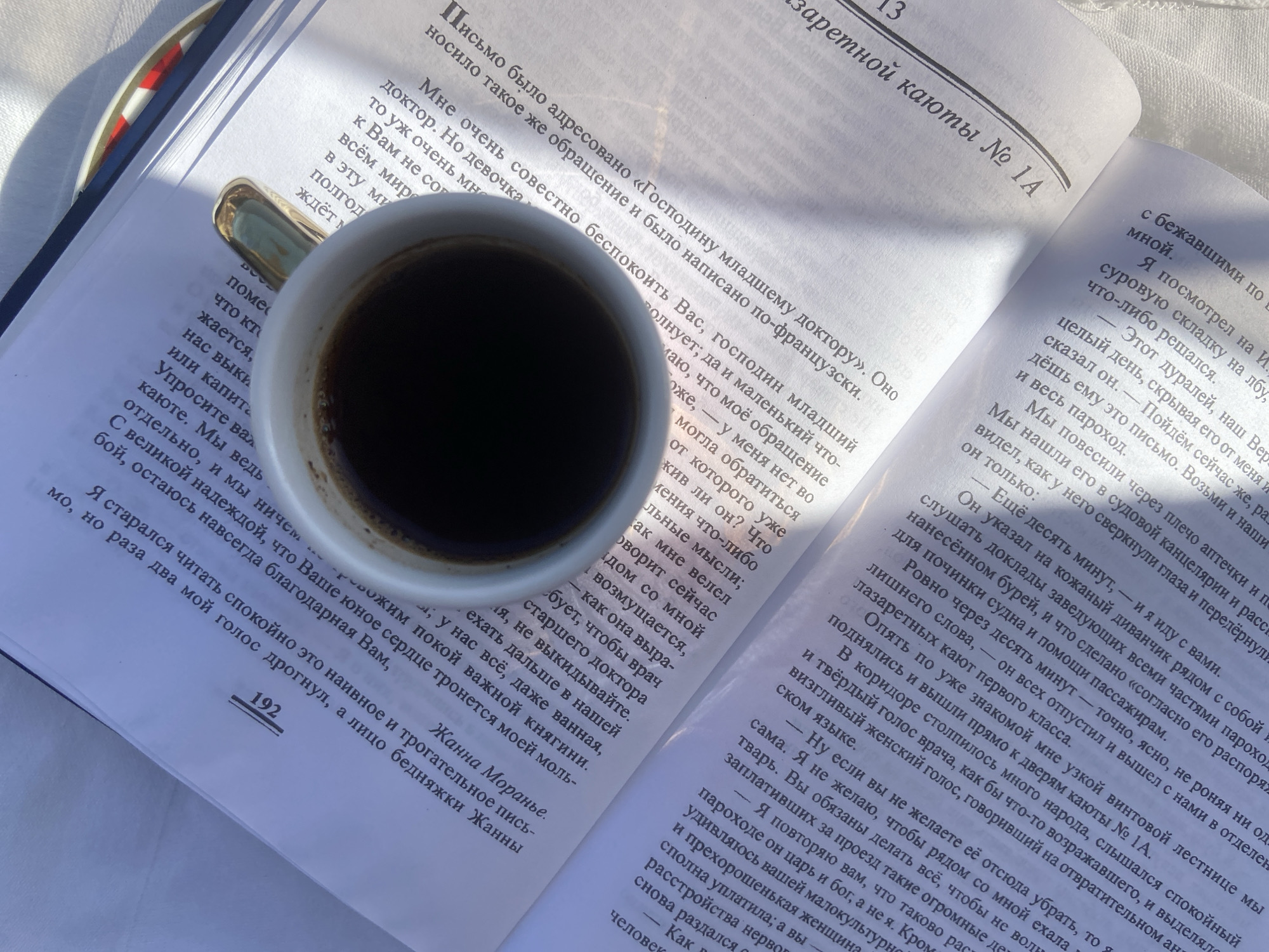 An overhead shot captures an open book with Cyrillic text and a white coffee cup with a gold handle placed on its pages. The book is open to a chapter titled "заретной каюты № 1А" (forbidden cabin No. 1A), with page number 192 visible at the bottom left. The coffee cup, filled with a dark beverage, rests on the left page.
Bright, natural sunlight illuminates the scene from the right, casting distinct shadows from the cup and the spine of the book onto the pages and the surface below. The surface beneath the book appears to be a light-colored, textured fabric.
Various passages of text are visible, predominantly in Russian. Notable phrases include: "Письмо было адресовано «Господину младшему доктору». Оно носило такое же обращение и было написано по-французски." (The letter was addressed to "Mr. Junior Doctor." It bore the same salutation and was written in French.) Another line reads: "Мне очень совестно беспокоить Вас, господин младший доктор." (I am very embarrassed to bother you, Mr. Junior Doctor.) Further down, the text includes "Я старался читать спокойно это наивное и трогательное письмо, но раза два мой голос дрогнул, а лицо бедняжки Жанны" (I tried to read this naive and touching letter calmly, but my voice trembled once or twice, and the face of poor Jeanne), followed by the name "Жанна Моранье" (Jeanne Moranière).