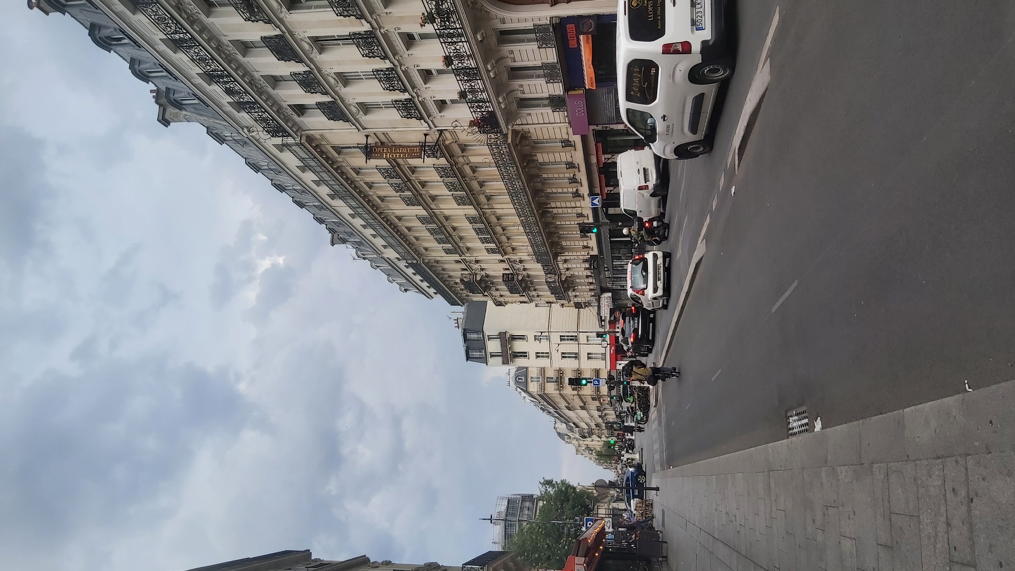 The image depicts a street scene in Paris, France, characterized by classic Haussmannian architecture. The buildings lining the street are tall and feature ornate wrought-iron balconies, mansard roofs, and numerous windows. The street itself is paved with asphalt and marked with white lines indicating lanes of traffic. On the right side of the street, a white van with the license plate "5023 LK 75" is prominently featured. Several cars and motorcycles are also visible in the road, some in motion and others stopped. A person is riding a bicycle on the left side of the street, near the sidewalk. The sidewalk itself is paved with cobblestones and lined with buildings that house shops and businesses, indicated by signs like "DOLLS" and "GOLETERIE." The sky is overcast with clouds, suggesting a typical Parisian day. Traffic lights are visible, with one showing a green signal. The overall impression is that of a bustling urban environment in Paris, likely during daytime.