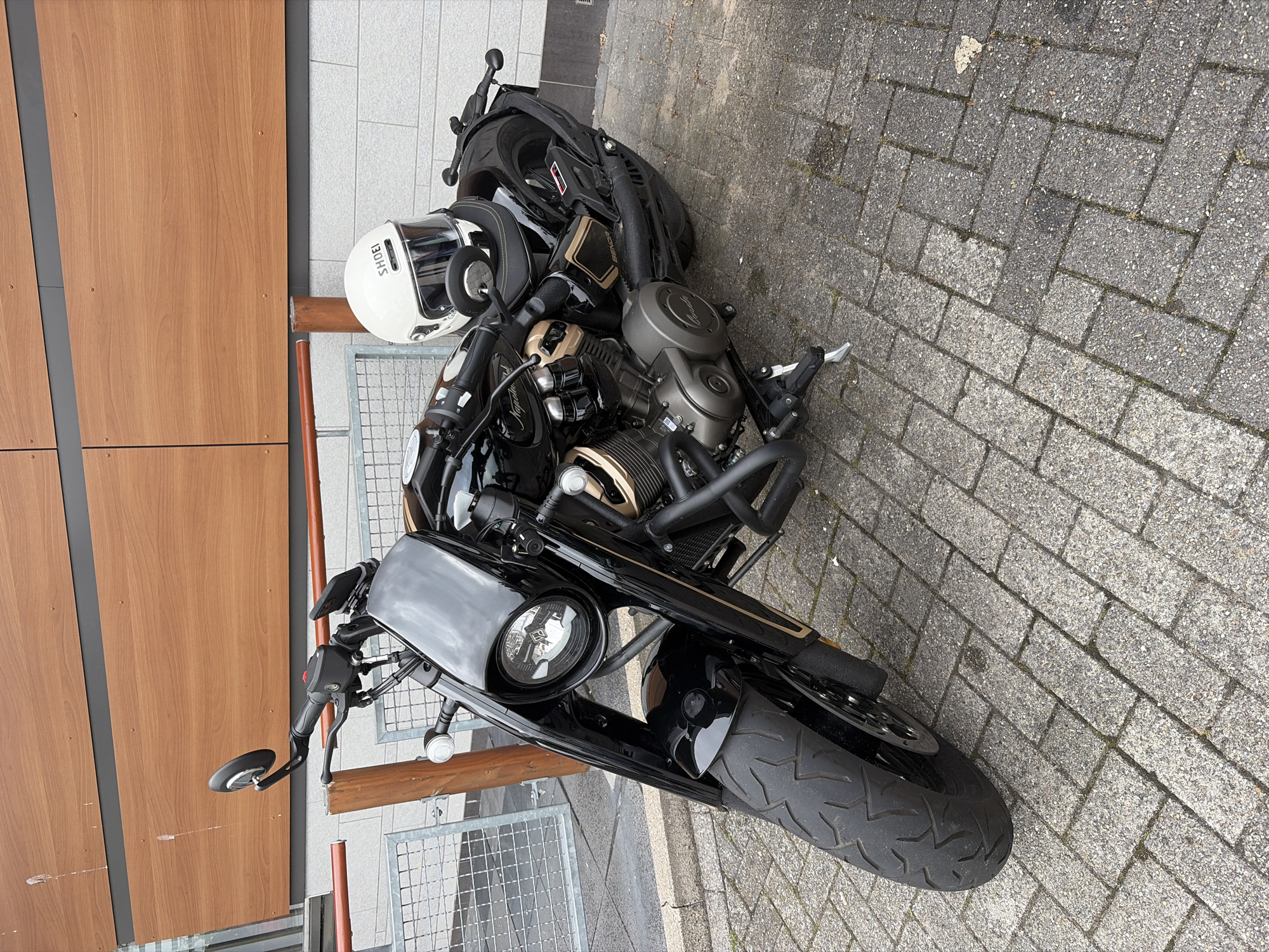 A black motorcycle with gold accents is parked on a brick-patterned sidewalk in front of a building with a wooden facade and a concrete wall. A white helmet with the brand "SHOEI" is placed on the motorcycle's seat. The motorcycle has a large front tire and appears to be in good condition. The environment suggests an urban or commercial setting. The building's design and materials do not provide specific clues to a particular location within Zoetermeer, Netherlands. There are no people visible in the image.