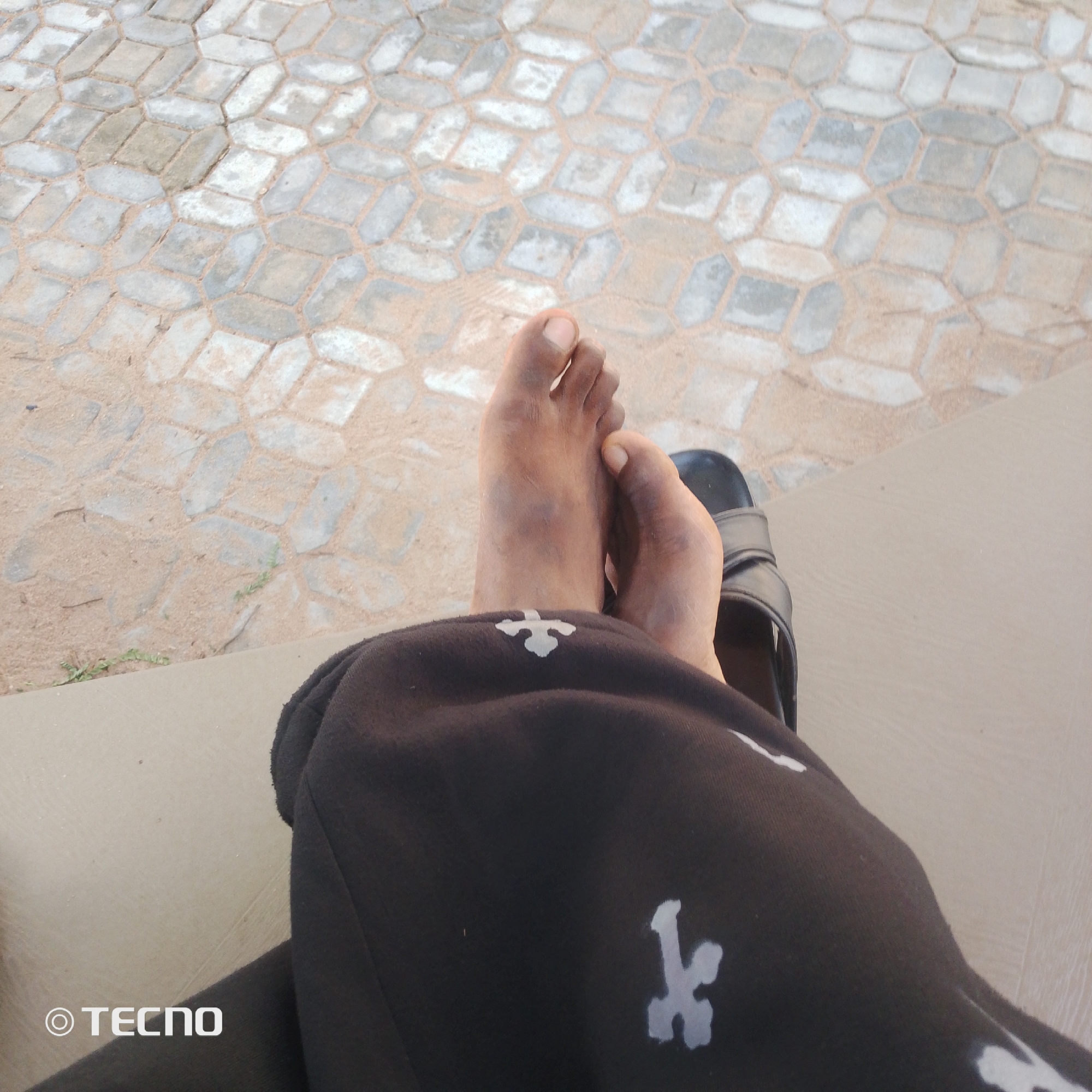 The image is a close-up shot of a person's feet, resting on a light-colored surface. The person is wearing black pants with a white pattern and black sandals. The background appears to be an outdoor paved area with interlocking concrete blocks. There is a visible "TECNO" watermark in the bottom left corner of the image. The scene is likely in Jalingo, Nigeria, given the watermark.