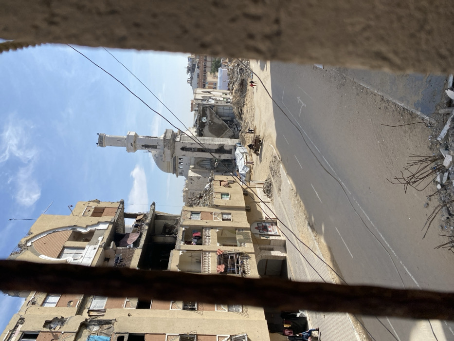 The image depicts a scene of extensive structural damage in Khan Yunis, Palestinian Territory, viewed from an elevated perspective. In the immediate foreground, exposed rebar and concrete debris are visible. A multi-story residential building dominates the left, featuring significant structural compromise, exposed interiors, shattered windows, and laundry draped on several balconies. A red and white circular sign is affixed to its lower level. An asphalt road with white lane markings spans the midground. On the left side of the road, a person sits or kneels on a paved area near the damaged building. To the right of the road, a mosque with a tall minaret and dome stands amid large piles of rubble, exhibiting significant structural damage to its lower sections and surrounding structures. Near the mosque, a three-wheeled motorized vehicle with one occupant is present, alongside two standing individuals. Further right, two additional people walk away from the mosque structure. Overhead utility wires intersect the clear blue sky with scattered white clouds.