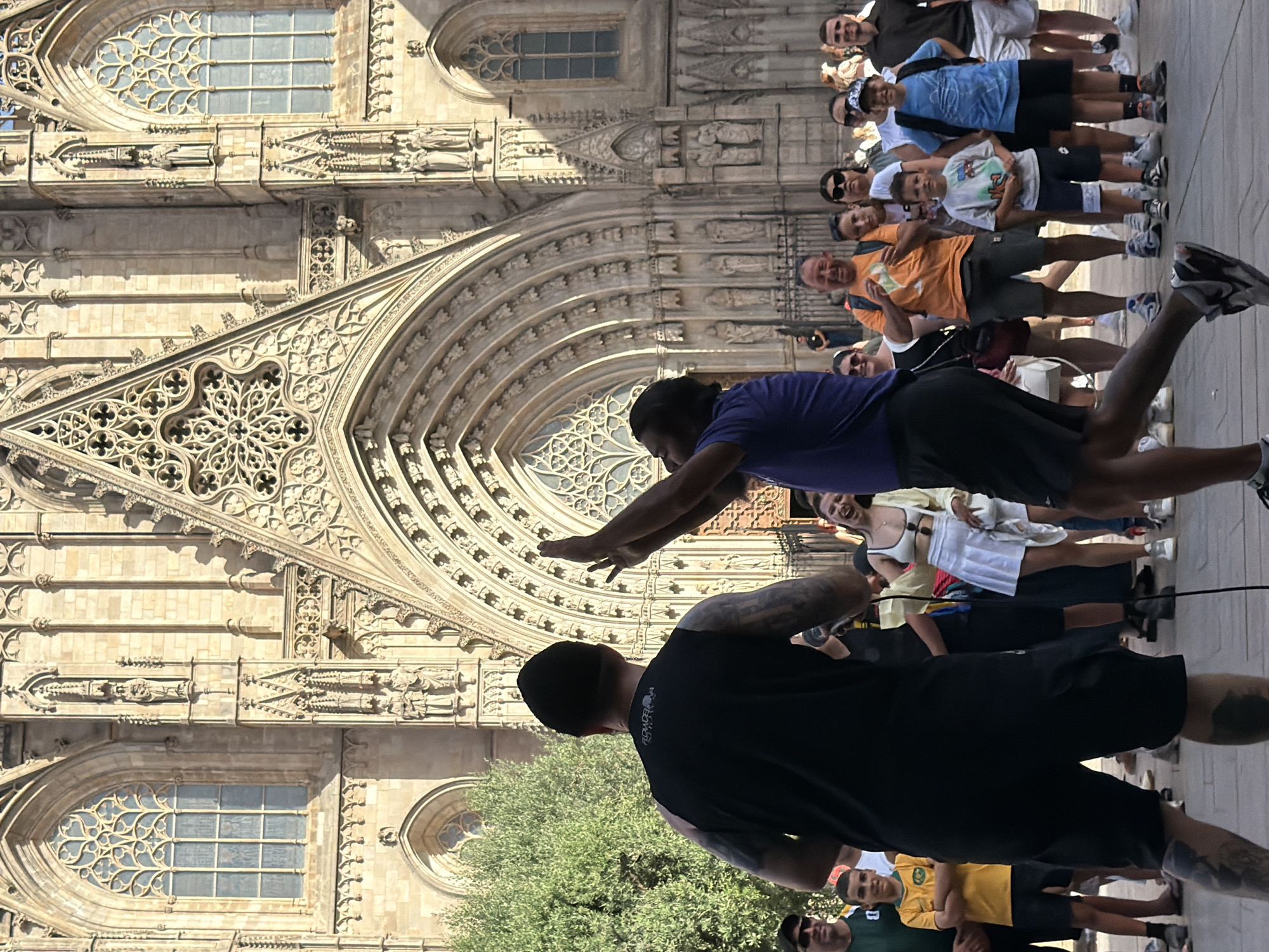 In Barcelona, Spain, a man in a black tank top and beanie, displaying tattoos on his arms, faces away from the camera and toward a crowd gathered in front of a large Gothic cathedral. He is holding a microphone. To his right, another man in a purple shirt with his back to the camera, also appears to be engaging with the crowd, raising his hands.
The crowd comprises a mix of adults and children, many looking towards the two men. Some individuals are smiling and appear to be enjoying the event. The architectural backdrop is a grand stone cathedral, characterized by intricate stonework, arched entrances, and large, ornate windows, typical of Gothic cathedrals found in European cities. The lighting suggests it is daytime, with the sun casting shadows. A lush green tree is visible to the left of the frame, providing a contrast to the stone architecture. The ground is paved with cobblestones.