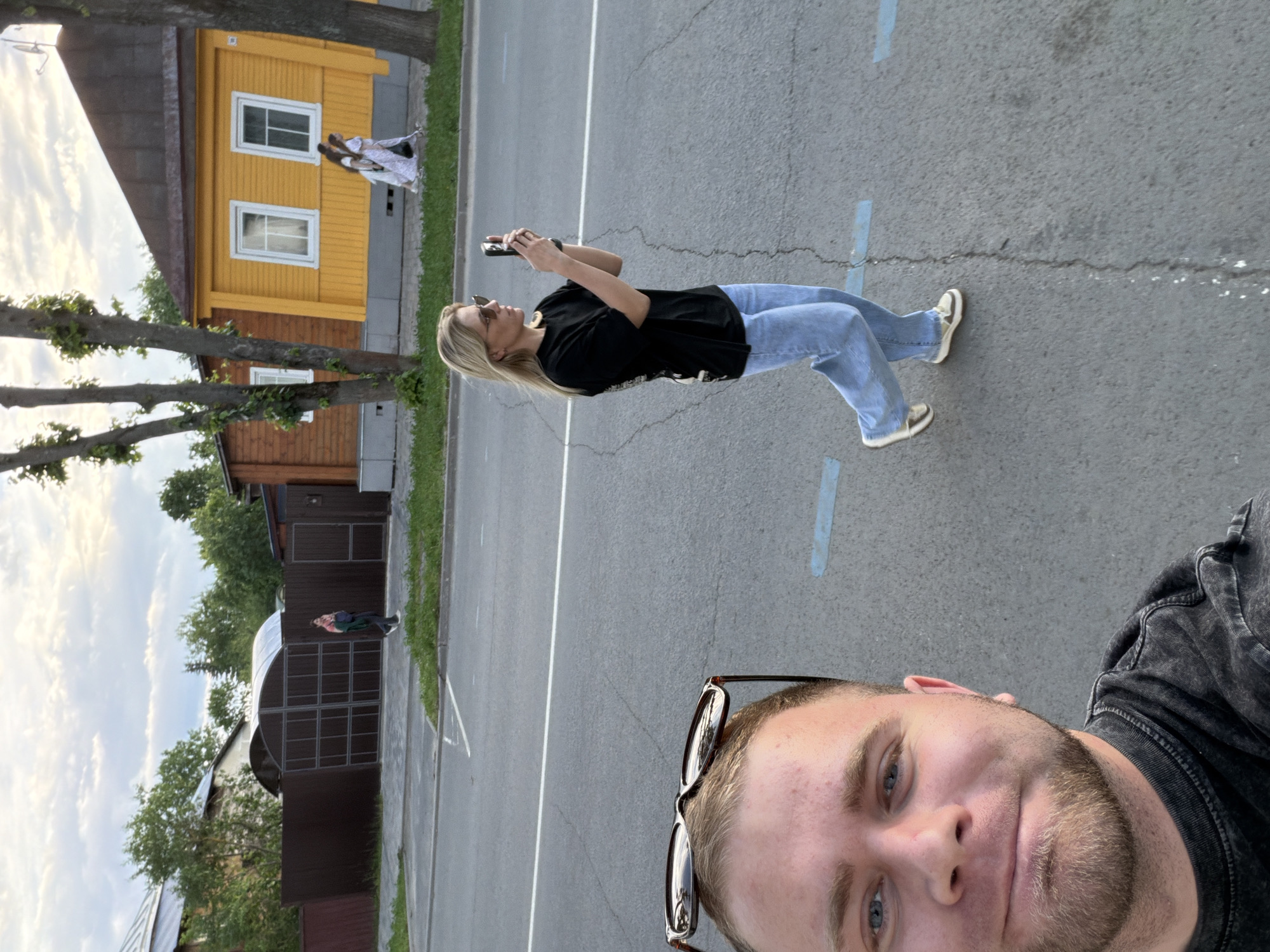 In Suzdal, Russia, a man is taking a selfie from a low angle, his face close to the camera. He has light skin, blue eyes, and a beard, and is wearing a dark t-shirt. His sunglasses are perched on top of his head. In the background, a woman with blonde hair is walking away from the camera, holding a smartphone to take a photo. She is wearing a black t-shirt and light blue jeans. The scene is set on a paved road with white and blue markings, with a grassy area and trees lining the street. A yellow building with white-framed windows stands to the right, and a dark brown fence encloses a property on the left. The sky above is cloudy.