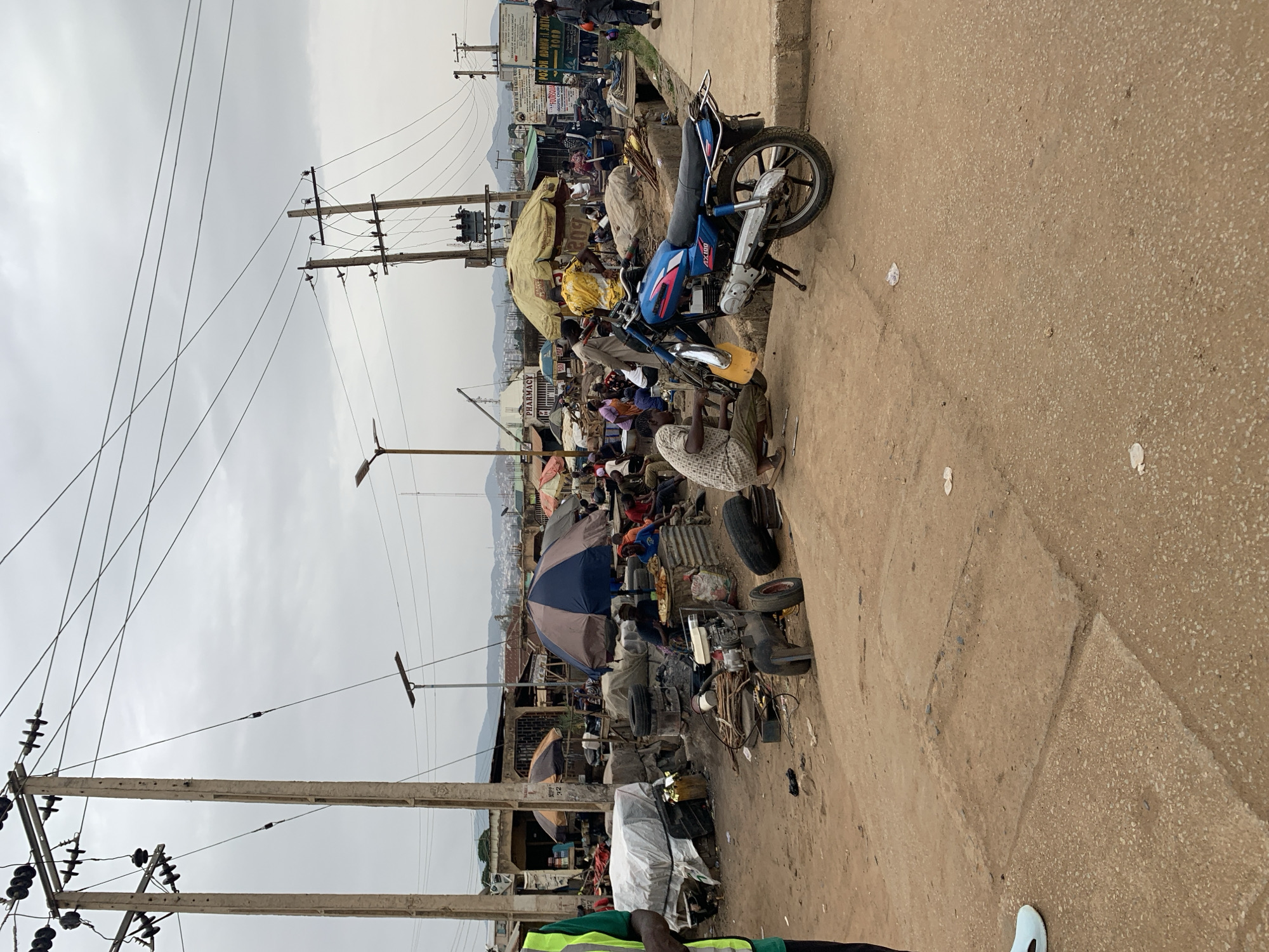 The image depicts a bustling street scene in Rafin Par, Nigeria. A blue and red motorcycle is parked on the side of a dirt road. The area is densely populated with people and covered stalls, suggesting a marketplace or commercial district. There are telephone poles with a tangle of wires overhead. The sky is overcast. People are walking or sitting amidst various objects, including tires, umbrellas, and miscellaneous goods.