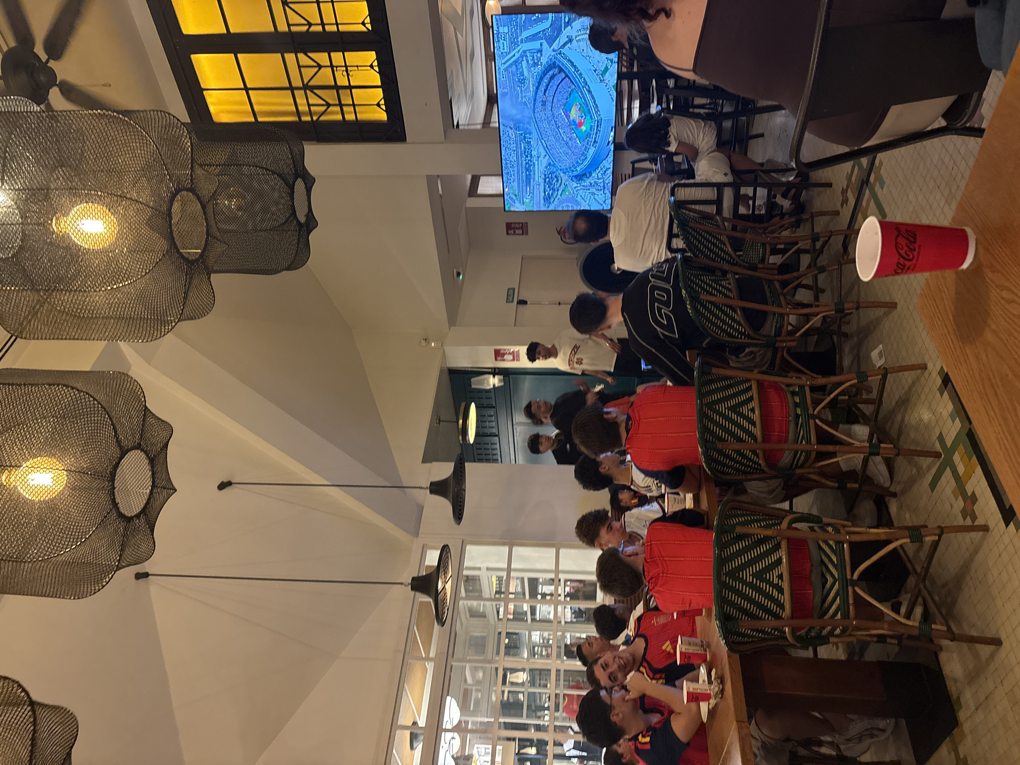 The image depicts a group of young men watching a large television screen displaying an aerial view of a stadium in a restaurant setting. Many of the men are wearing red and yellow jerseys, suggesting they are fans of a sports team, possibly Spain's national football team, given the colors. The restaurant has a modern industrial design with large mesh pendant lights and tall windows. The floor is tiled with a mosaic pattern. Several people are seated at wooden tables and chairs, some with Coca-Cola cups. The atmosphere appears casual and engaging, with the individuals focused on the screen. One person in the foreground is wearing a black shirt with white lettering that reads "COL". The presence of a "SALIDA" (exit) sign and fire extinguisher suggests this is a public establishment. The scene could be set in a restaurant or bar in the city of València, Spain, given the likely context of watching a sporting event with Spanish team colors.