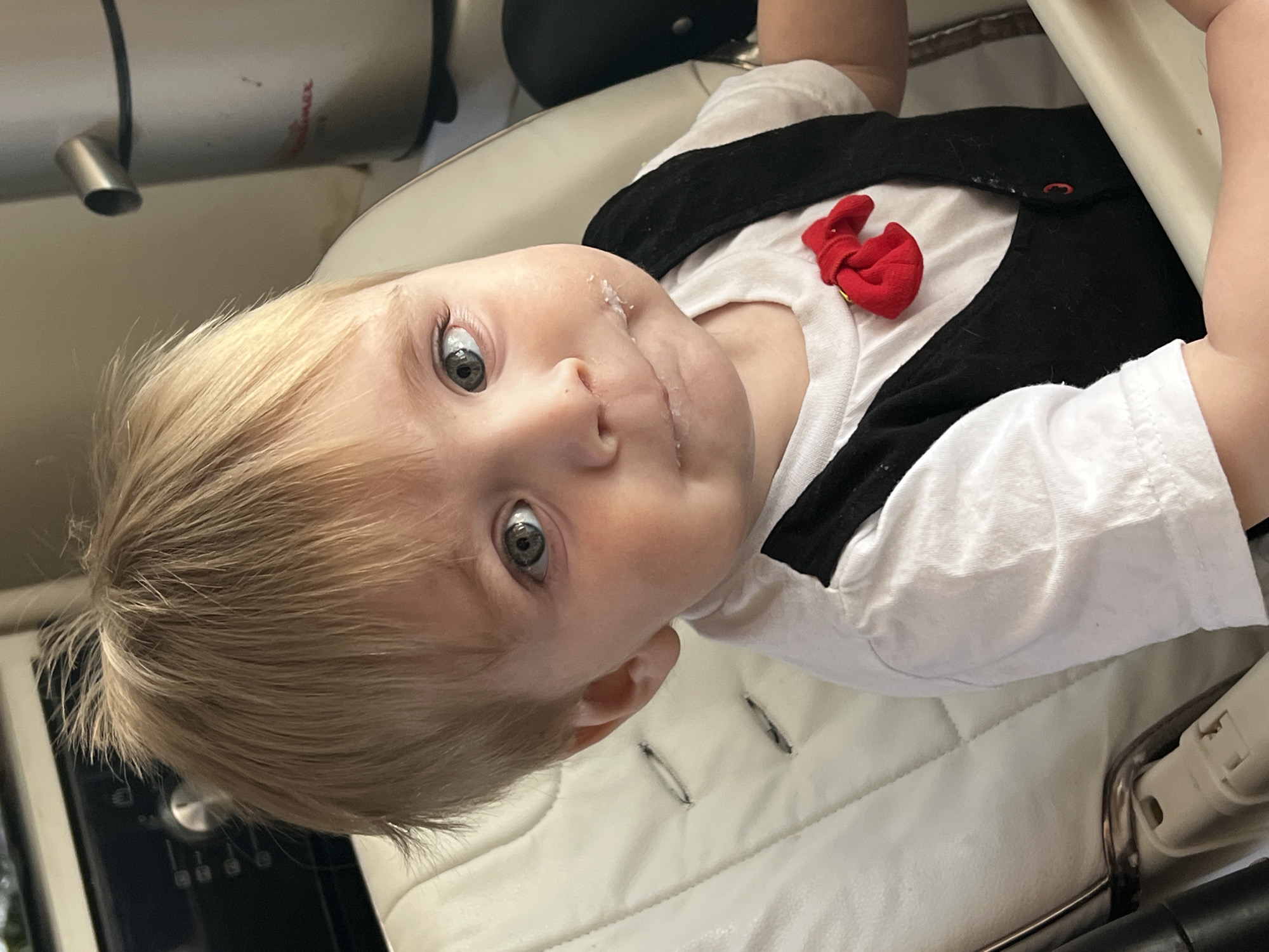 A toddler with blond hair and gray eyes is looking up at the camera. The toddler is wearing a white t-shirt with a black vest and a red bow tie. The toddler has white residue on their face. The toddler is sitting in a white padded seat with metal supports. A stainless steel cylindrical object is visible in the upper left corner of the image, along with a black object above the child's head. The location and city of Noviy, Russia, are not determinable from this image.