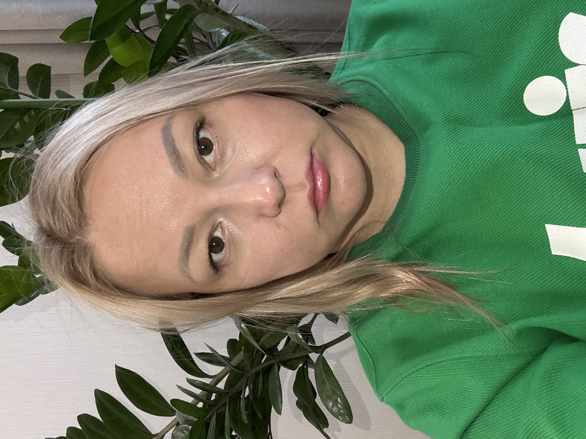A woman with blonde hair is positioned slightly off-center, facing the camera with a direct gaze. She has fair skin, brown eyes, and pink lips. She wears a bright green sweatshirt, part of which is visible in the upper right corner of the frame.
The background includes a plant with green leaves, partially framing the woman's face and upper body on both sides. A white surface or wall is visible to the left of the plant.
There is no information indicating a location in Izhevsk, Russia. The image is a close-up portrait and does not contain geographic or contextual clues that would enable location identification.