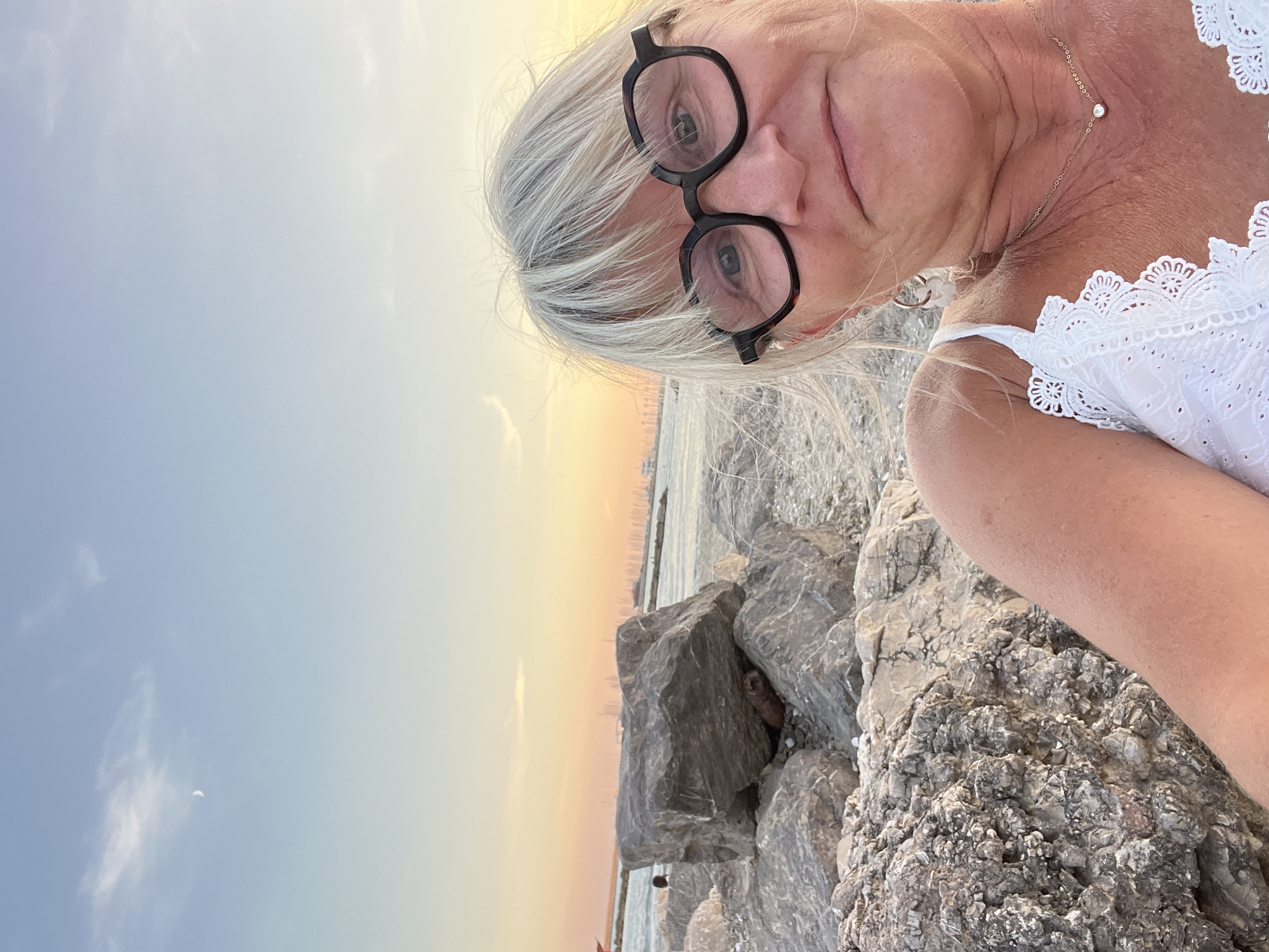 A woman with blonde hair and glasses is taking a selfie by the sea. She is wearing a white top and a necklace. She is surrounded by rocks. The sky is a pale orange and blue, with a crescent moon visible. The ocean is in the background, with a city skyline in the distance. The location is Palavas-les-Flots, France.