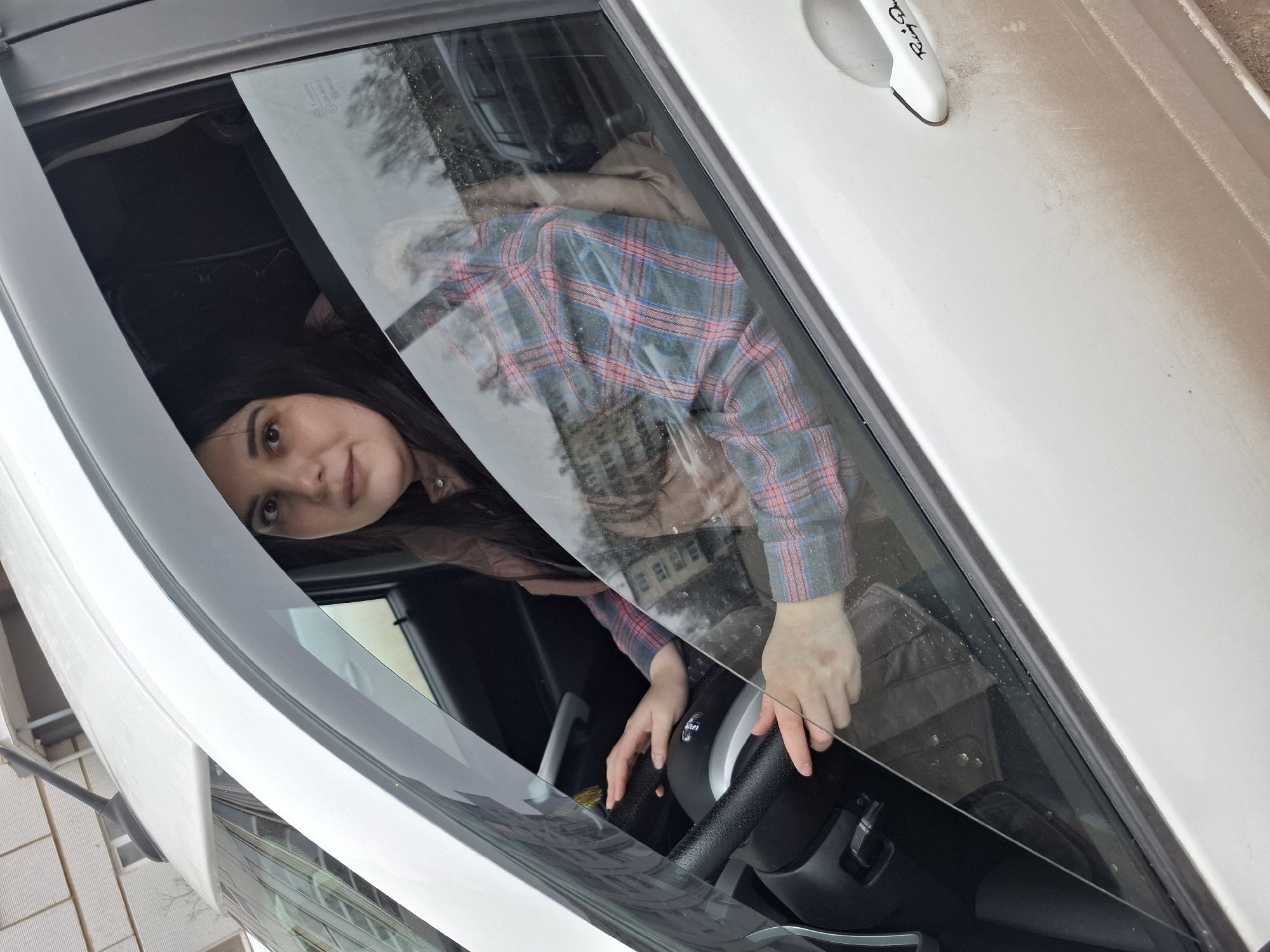 A young woman with long dark hair is captured sitting in the driver's seat of a white car, looking directly at the camera through the partially open window. She wears a plaid shirt featuring blue, red, and white checks over a light brown jacket. Her hands rest on the steering wheel, and she has a neutral to slightly smiling expression.
The car's exterior is white, with a noticeable layer of dust or dirt on the door surface. Reflections in the window reveal multi-story buildings, suggesting an urban or residential outdoor environment, consistent with a location in Chaikovsky, Russia. The natural lighting indicates it is daytime, possibly under an overcast sky, and there are small spots on the window glass, which could be raindrops or dirt.
Faint text is visible on the car's white door handle, appearing to read "Rally Das" or a similar phrase. The car's interior visible through the window is dark. The image is taken at an oblique, rotated angle.
