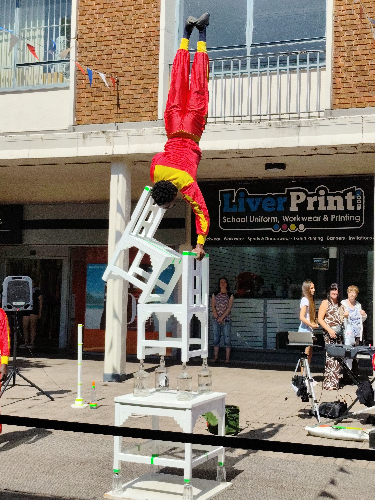 The image depicts an outdoor street performance in Knowsley, United Kingdom. A male performer, dressed in bright red and yellow trousers and top, is balanced upside down on a stack of white chairs. The chairs are arranged precariously, with some supported by glass bottles and others resting on upturned glasses. In the background, a building with a brick facade and large windows is visible, with a sign for "LiverPrint.com" advertising "School Uniform, Workwear & Printing." Several spectators, including women of various ages, are observing the performance. The ground is paved with paving stones, and a black rope barrier separates the performance area from the public. A speaker on a stand and musical equipment are also present, suggesting a performance involving music or amplification. The overall scene suggests a public event or festival taking place during daylight hours.