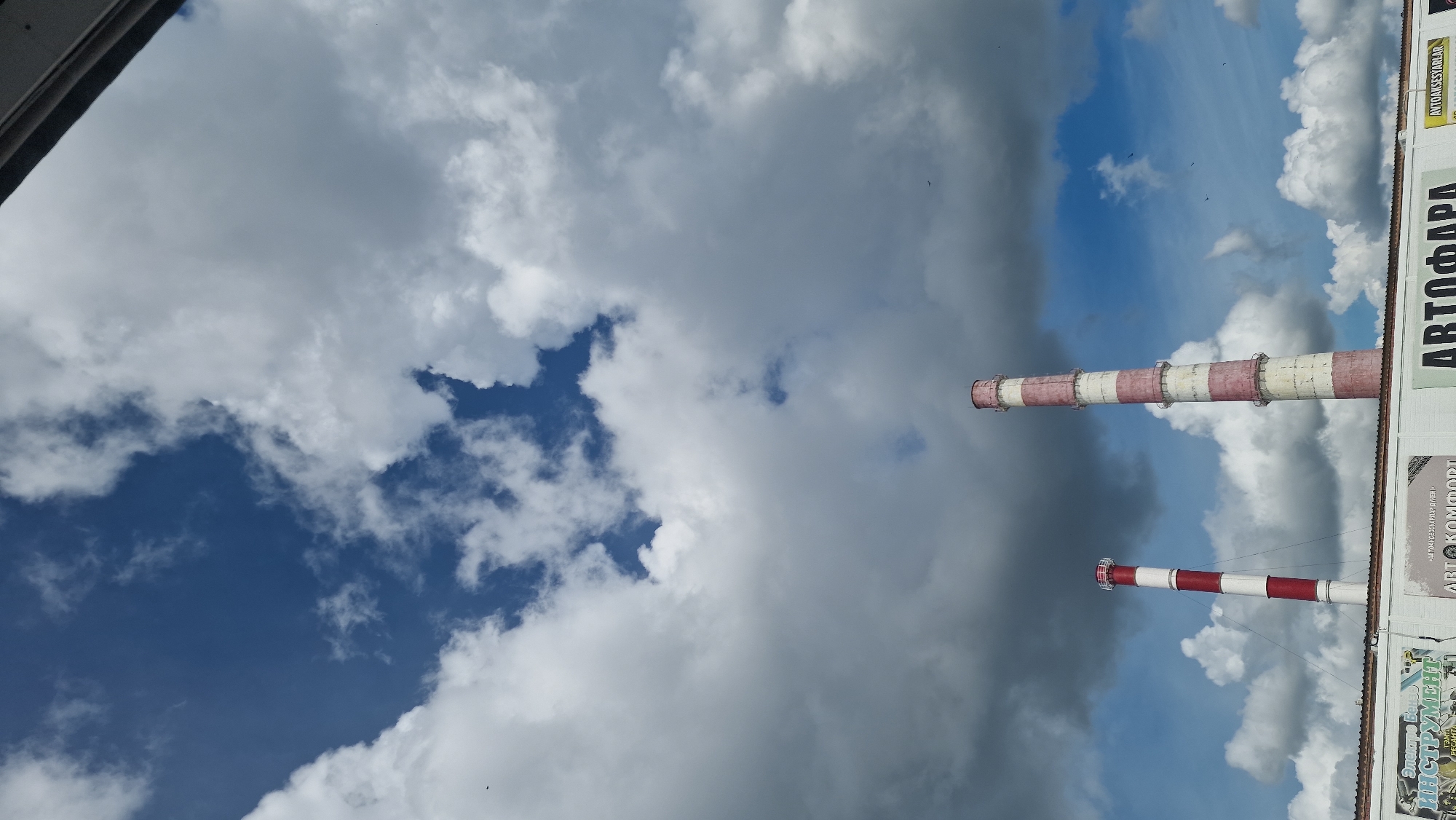 The image depicts two industrial chimneys, one taller and more prominent on the right, the other shorter and to the left. Both chimneys are striped with red and white sections and extend upwards into a sky filled with dramatic clouds, ranging from dark grey to bright white, interspersed with patches of blue sky. The chimneys appear to be part of an industrial facility, as suggested by the rooftop structure visible at the bottom of the frame, which features various signs. One sign is partially visible and appears to advertise "Автокомфорт" (Avtokomfort), indicating a business related to automotive comfort or services. The overall scene suggests an industrial or commercial area under a cloudy sky.