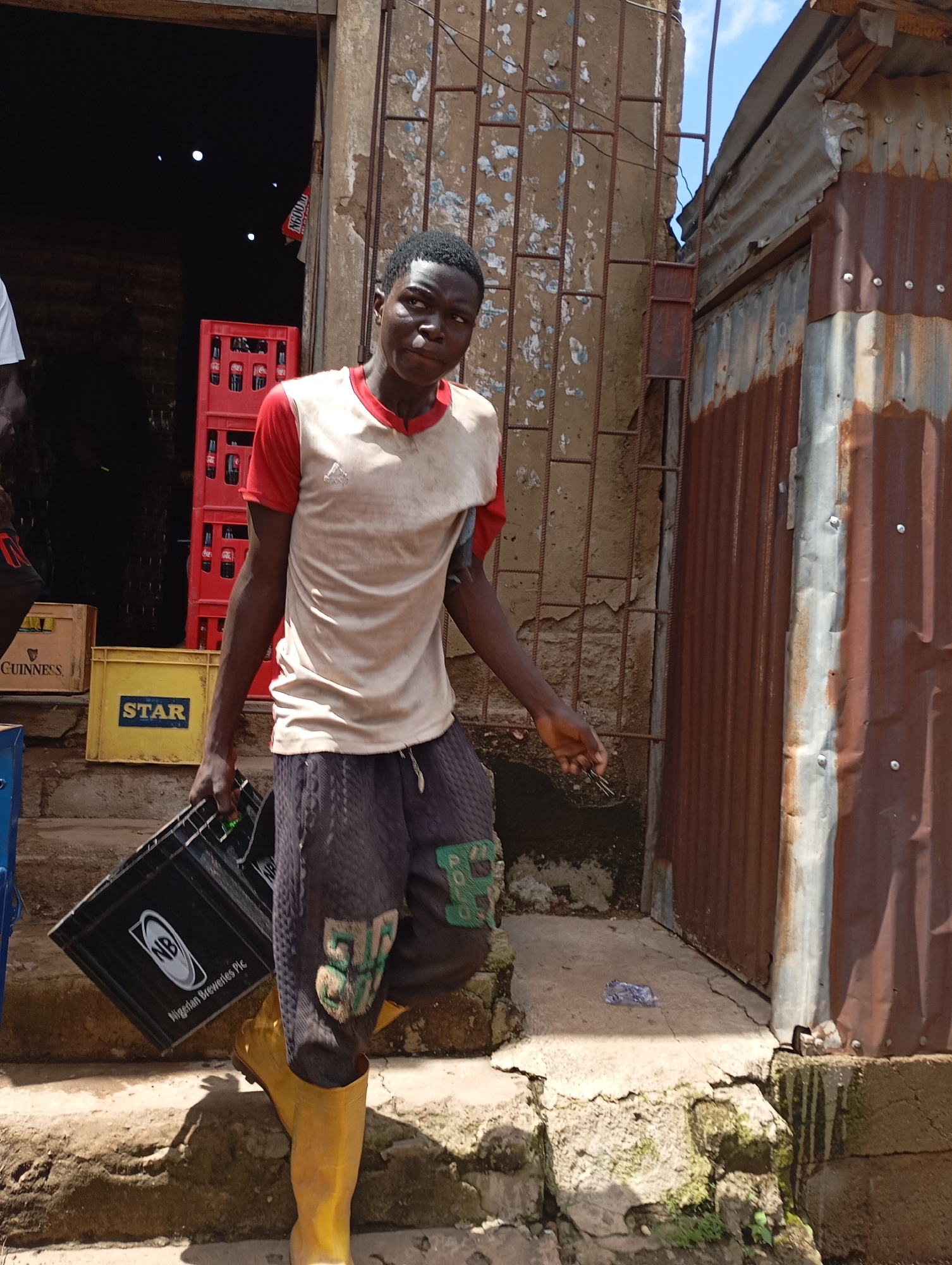 A young Black male in Shendam, Nigeria, wearing a red and white t-shirt, grey patterned shorts, and yellow rubber boots, carries a black crate with the "Nigerian Breweries Plc" logo. He is walking down concrete steps towards a rustic building with corrugated metal siding and a barred window. In the background, red crates stacked with bottles, likely beverages, are visible inside a dark doorway. The ground is cracked concrete, and the environment appears weathered and urban. The man is looking towards the camera with a neutral expression.