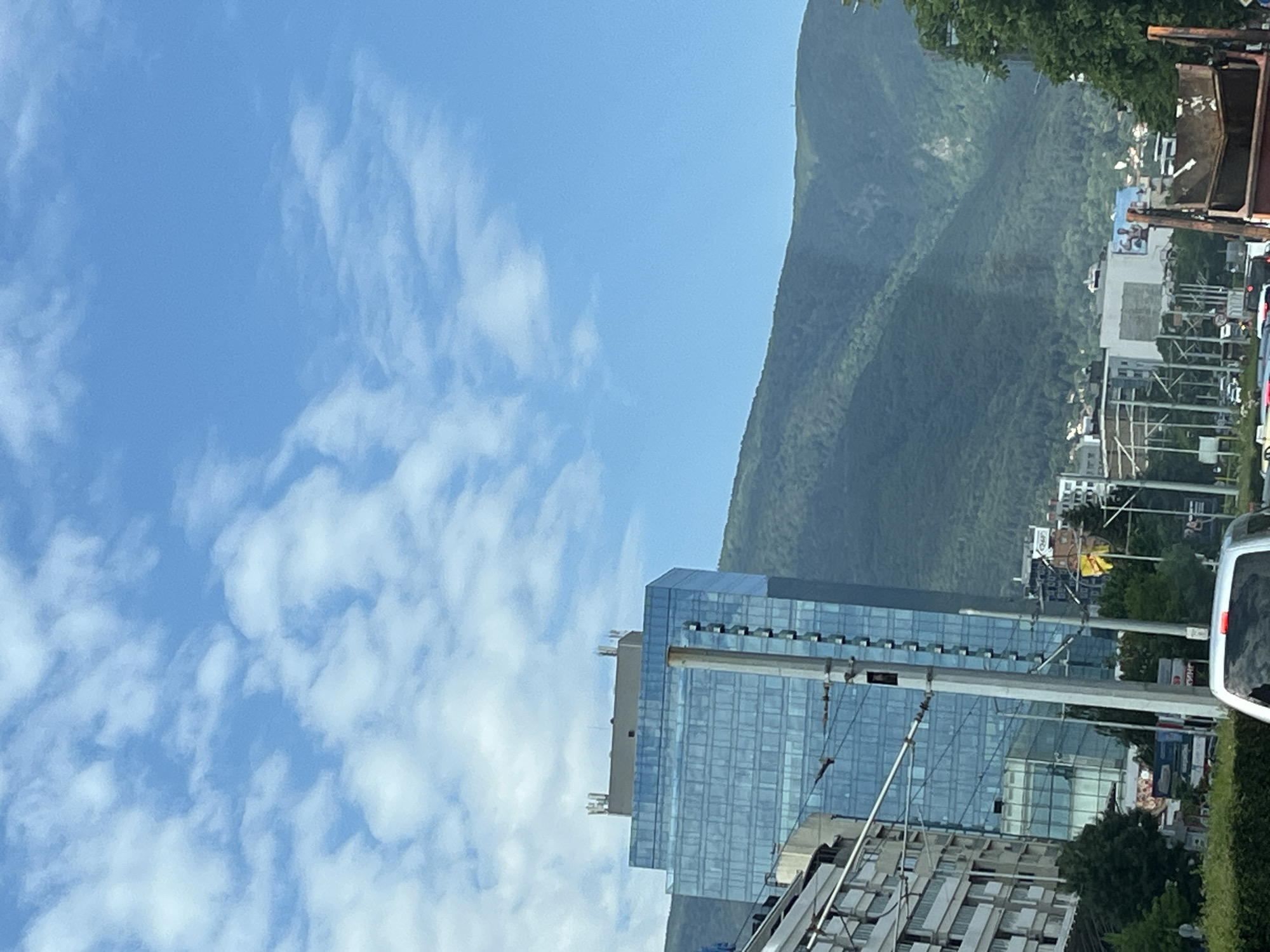 A low-angle medium shot captures a section of a city against a backdrop of lush green mountains under a bright, cloudy sky. In the foreground, a modern glass-fronted skyscraper with a beige top stands tall, reflecting the sky. To its left, a section of a white building with multiple windows is visible. Several utility poles with wires crisscross the frame in front of the skyscraper, partially obscuring it. In the mid-ground, more buildings and trees are seen, with a yellow flag flying prominently among them. The background is dominated by a vast, verdant mountain range. The scene appears to be a street-level view, possibly from a vehicle, with traffic and foliage in the lower portion of the image.