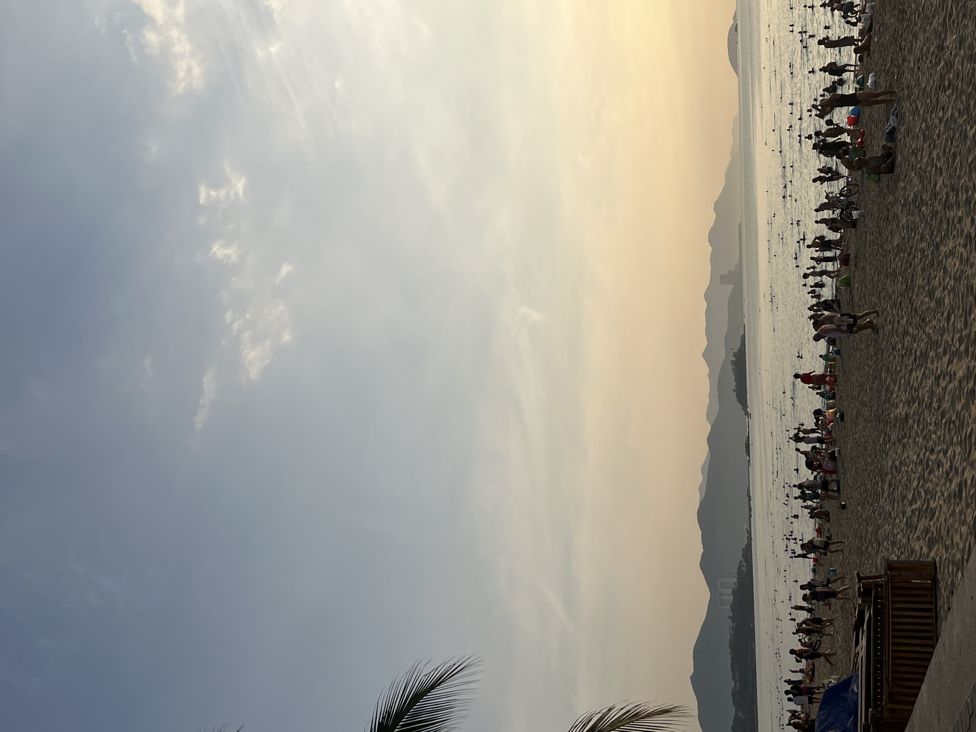 The scene shows Nha Trang beach in Nha Trang Ward, Vietnam, at what appears to be either sunrise or sunset, given the diffused yellow-orange light. A long stretch of sandy beach runs along the right side of the image, populated by many people. Individuals are observed near the water, seemingly engaging in swimming or wading. Dark figures dispersed throughout the water suggest the presence of buoys or other floating objects. The background features a mountainous landscape partially obscured by haze, indicative of distance. The sky is partly cloudy with a color gradient shifting from gray-blue to yellow, corresponding with the light. Parts of the palm tree are also visible in the bottom-left of the frame.