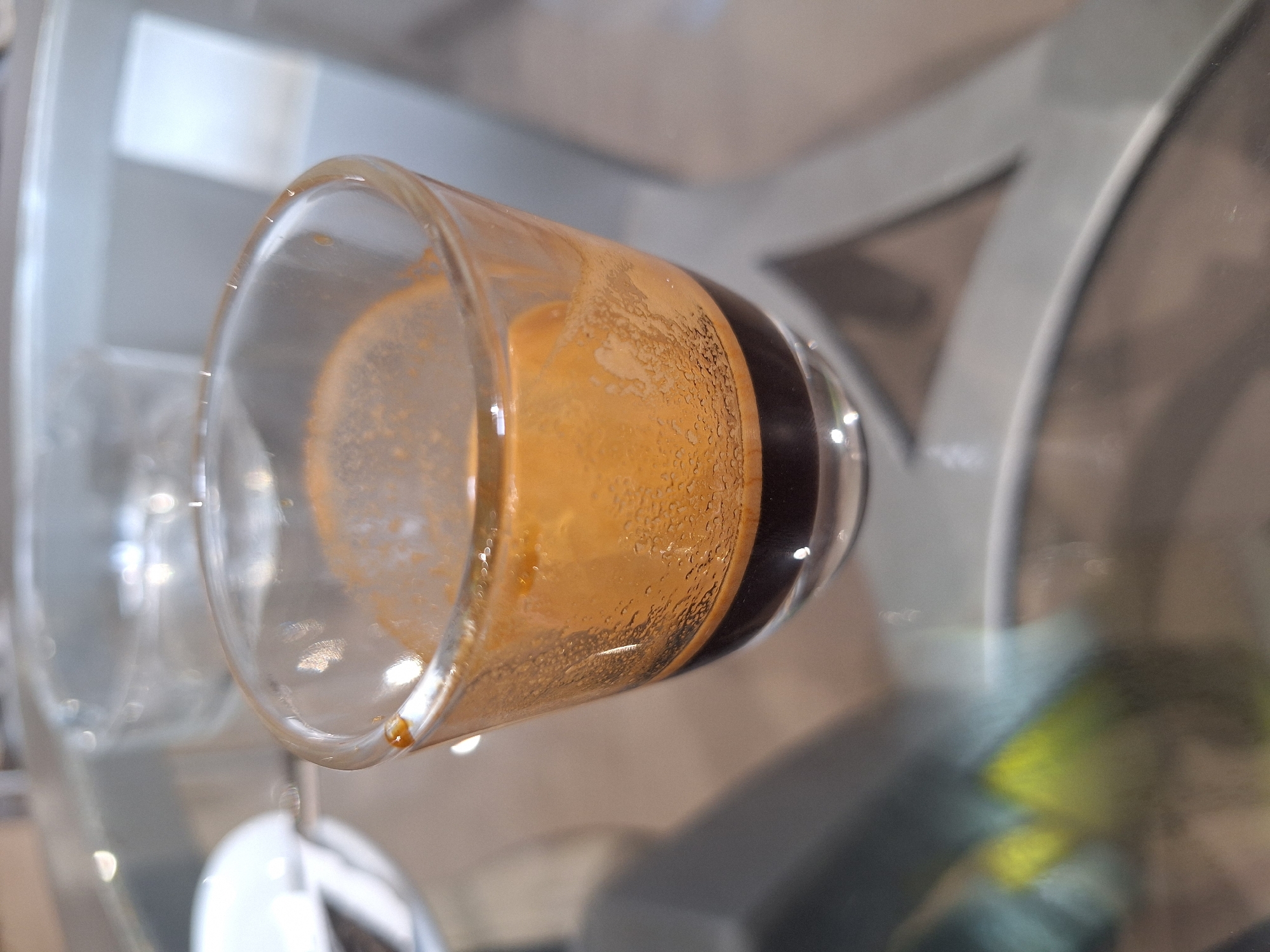 This image, captured in Casablanca, Morocco, features a single espresso shot presented in a small, clear, footed glass. The espresso exhibits a dark brown liquid base beneath a substantial layer of golden-brown crema, which displays numerous small bubbles clinging to the interior sides of the glass. A distinct drip of coffee is visible on the upper left exterior rim of the glass. The glass is positioned on a reflective, transparent glass tabletop, which shows reflections of light sources and blurred elements from the environment. Through the glass table, the blurred support structure of the table, appearing light-colored and geometric (possibly X-shaped), is discernible. The background is heavily out of focus, indicating an indoor setting, with blurry shapes suggesting another clear glass and a white object (possibly a saucer) on the left. No individuals are present in the frame.