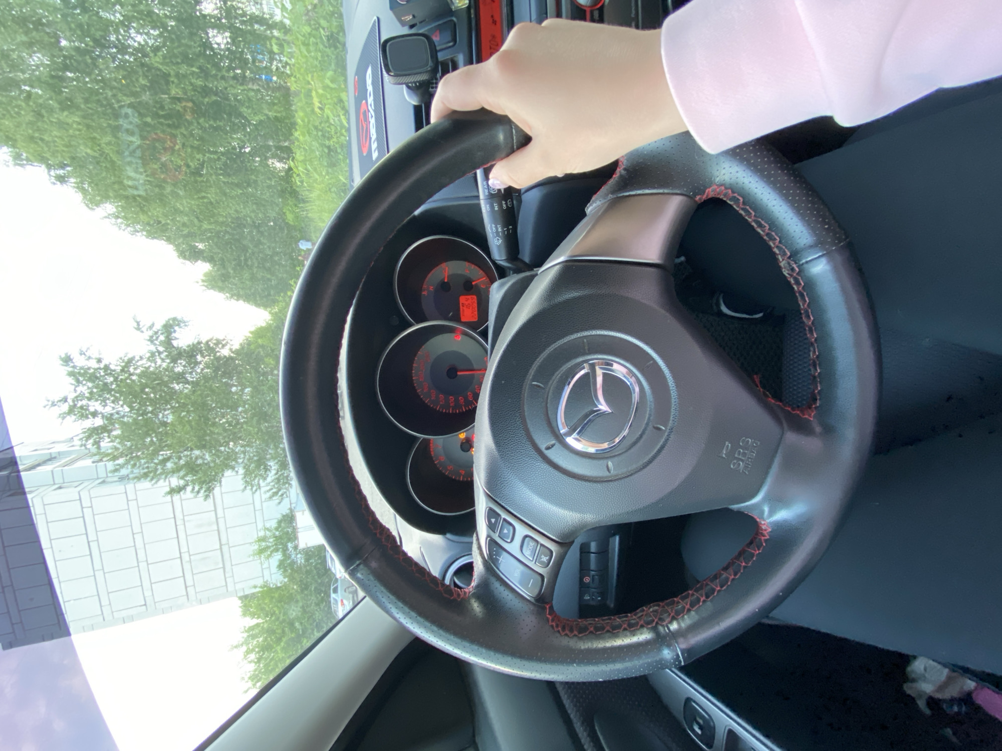 The image shows the interior of a Mazda car, focusing on the steering wheel and dashboard. A hand is holding the steering wheel. The dashboard instruments are visible, including the speedometer and tachometer. Outside the car window, greenery and a building are visible, suggesting an urban environment. Without more detail, I can not provide information about the location in the city of Chaikovsky, Russia.