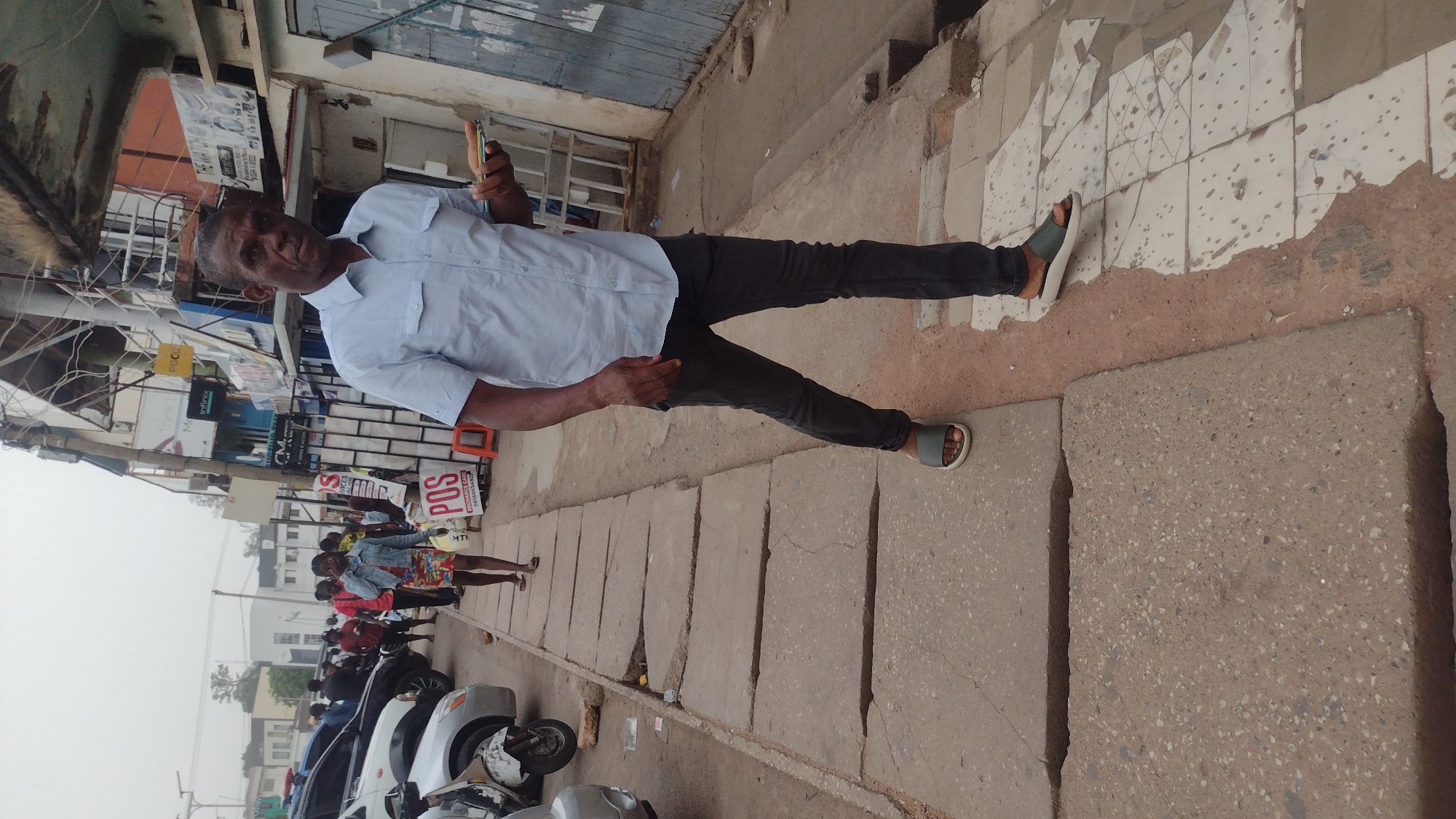An adult male stands on a city street in Jos, Nigeria. He wears a light-blue short-sleeved button-down shirt, dark pants, and sandals. He holds a mobile phone in his right hand. The street features a concrete sidewalk and curb with rectangular covers lining the edge. Behind the man is a building with shops. Several people walk in the background along the sidewalk near parked motorcycles and cars. Signage indicating services such as POS and MTN is visible.