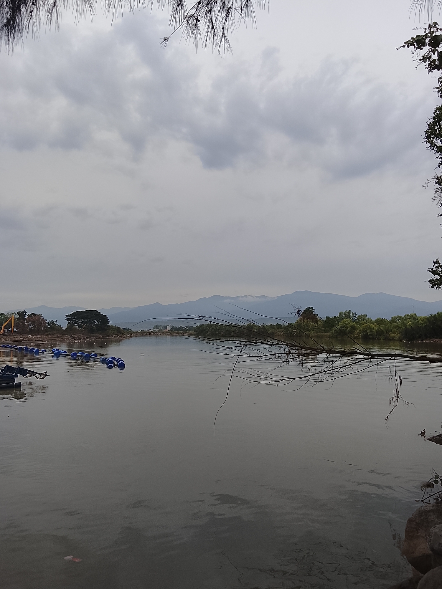 The image depicts a serene natural landscape with a body of water in the foreground, reflecting the overcast sky. In the distance, a mountain range stretches across the horizon, its peaks softened by the atmospheric haze. The water's surface is disturbed by gentle ripples, and a few floating buoys are visible on the left side, suggesting some form of aquatic activity or construction. A few trees and some vegetation are scattered along the banks, adding to the natural beauty of the scene. The overall impression is one of tranquility and natural splendor. This scene is believed to be in the city of Reudeuep, Indonesia.
