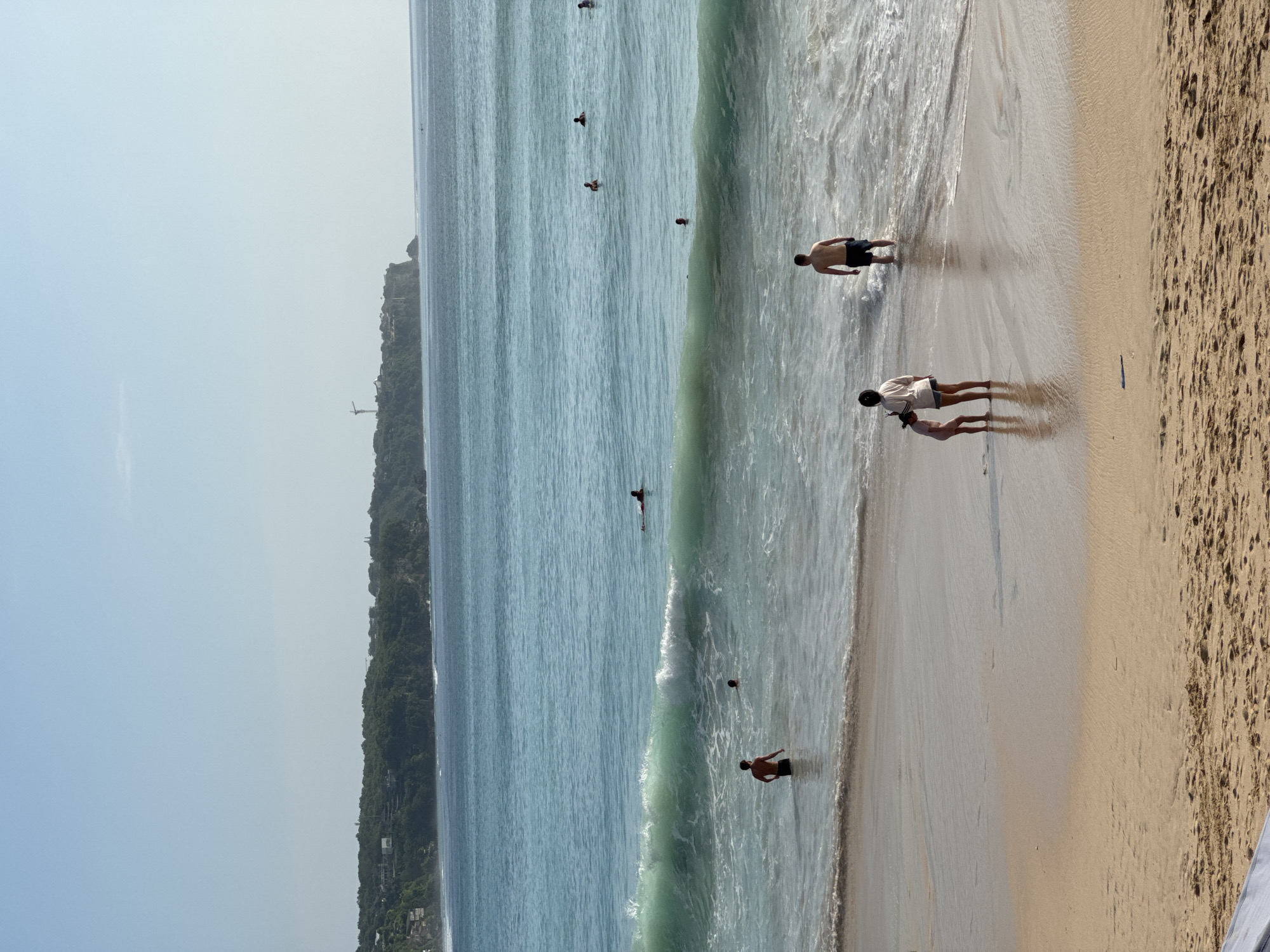 The image captures a vibrant, sunny beach scene in Kuta Selatan, Indonesia, viewed from a slightly elevated vantage point. The expansive turquoise ocean meets a wide, golden-brown sandy shore, backed by a prominent, lush green landmass under a clear, light blue sky.
Several people are scattered across the scene, engaging in various leisure activities. On the wet sand in the foreground, a shirtless man in dark shorts stands at the water's edge, looking towards the ocean. Further up the beach, an adult in a light shirt and dark shorts stands alongside a child, both casting distinct shadows on the sand, suggesting a bright midday or early afternoon sun. In the ocean, multiple individuals are enjoying the water: one person stands in the shallow waves, while others are swimming or wading further out, with one person appearing to be on a board.
The notable details include the calm, clear ocean with gentle breaking waves near the shore. The extensive landmass in the background is a verdant, hilly peninsula or cliff, densely covered in tropical vegetation. Visible on this landform are some man-made structures, including what appears to be an elevator or funicular system descending the cliff face, and a distant, slender structure resembling a lighthouse or monument on the highest point. The sand transitions from wet and dark near the waterline to drier and lighter further up, showing some footprints and minor debris. The overall impression is one of relaxation and recreation on a beautiful, clear-weather day. There is no visible text in the image.