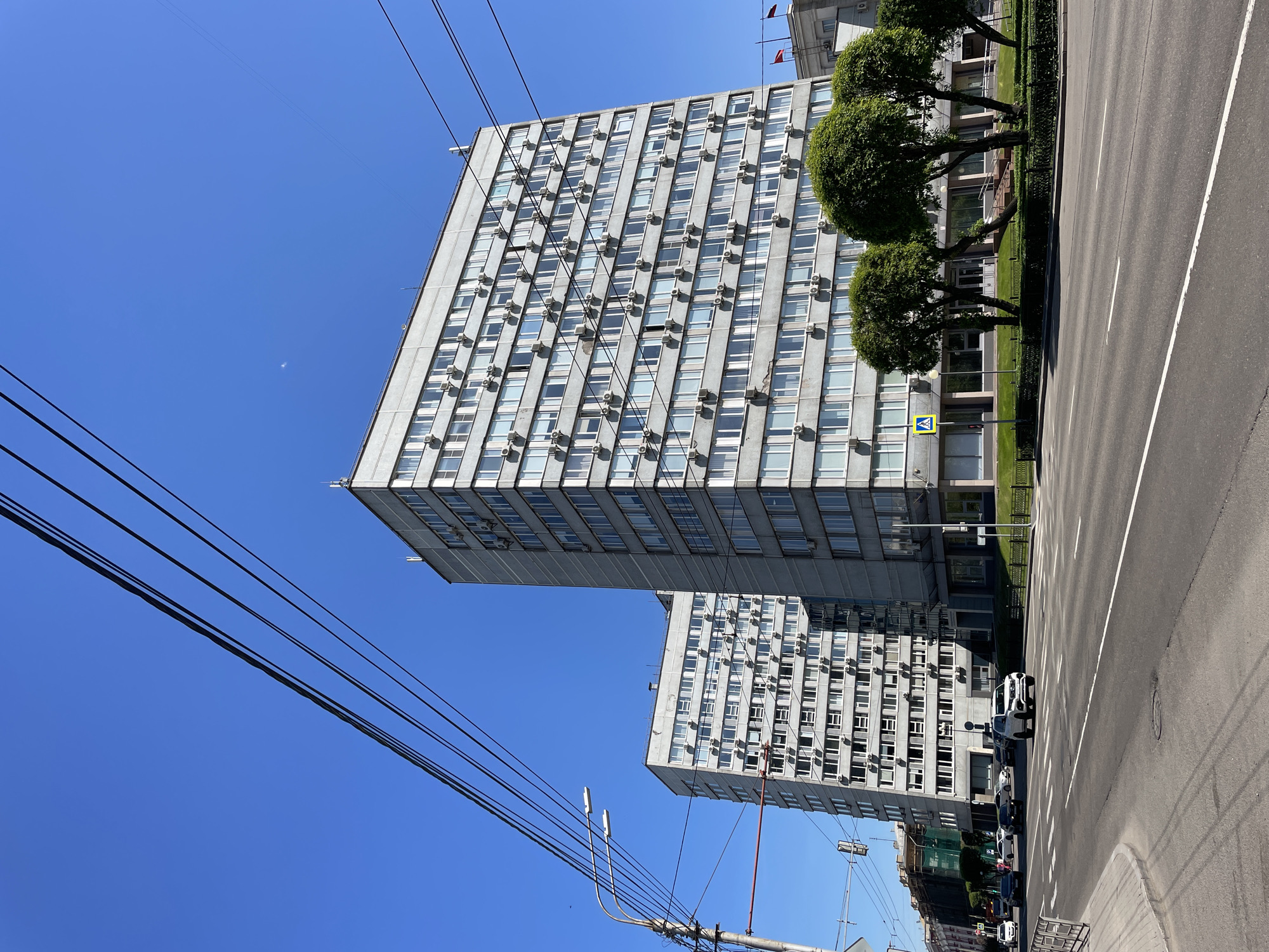 This media file captures an urban street scene on a bright, clear day, likely in Krasnoyarsk, Russia. The image is oriented vertically, with the top of the photo showing what would be the right side of a conventionally oriented street view.
Two large, multi-story buildings, constructed from light grey, utilitarian-style concrete, dominate the left side of the frame. The facade of the prominent front building is covered with a grid of numerous windows, almost all of which have an external air conditioning unit mounted beneath them. A second, similar building is partially visible further back.
To the right of the buildings, a wide asphalt road with white dashed and solid lane markings stretches into the distance. A strip of green lawn, featuring several neatly trimmed, spherical trees, separates the buildings from the roadway. A yellow triangular pedestrian crossing sign is visible on this green strip near the front building. A few parked cars can be seen along the road further away.
The upper-left portion of the image is filled with a vast expanse of clear, bright blue sky, crisscrossed by multiple black power lines or trolleybus wires. A faint, crescent moon is discernible in the clear sky.
The scene depicts a typical urban environment with no specific activity or event taking place. The bright sunlight and absence of clouds indicate good weather, likely mid-day to early afternoon. The visible text is limited to the universal pedestrian crossing symbol on the yellow sign. The architecture and infrastructure are characteristic of a city in Russia.