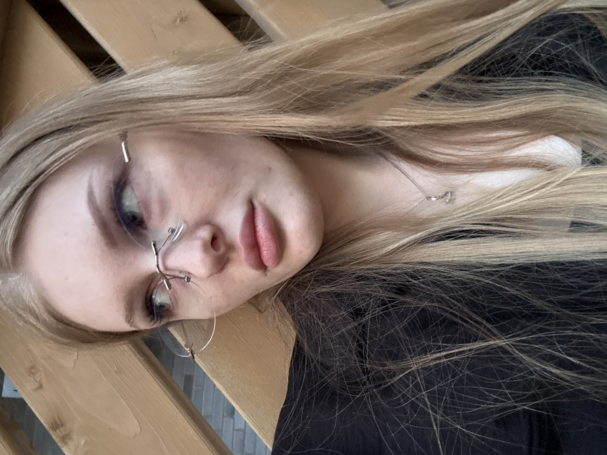 A young adult female with light blonde hair is photographed at a slight angle, partially facing the camera. She is wearing rimless glasses with a distinctive bridge design and hoop earrings. She has fair skin and is wearing makeup, including eyeliner. A delicate silver necklace is visible on her neck. She is wearing a dark-colored shirt or dress. The background includes a wooden structure, possibly a railing or fence. The location is Perm, Russia.