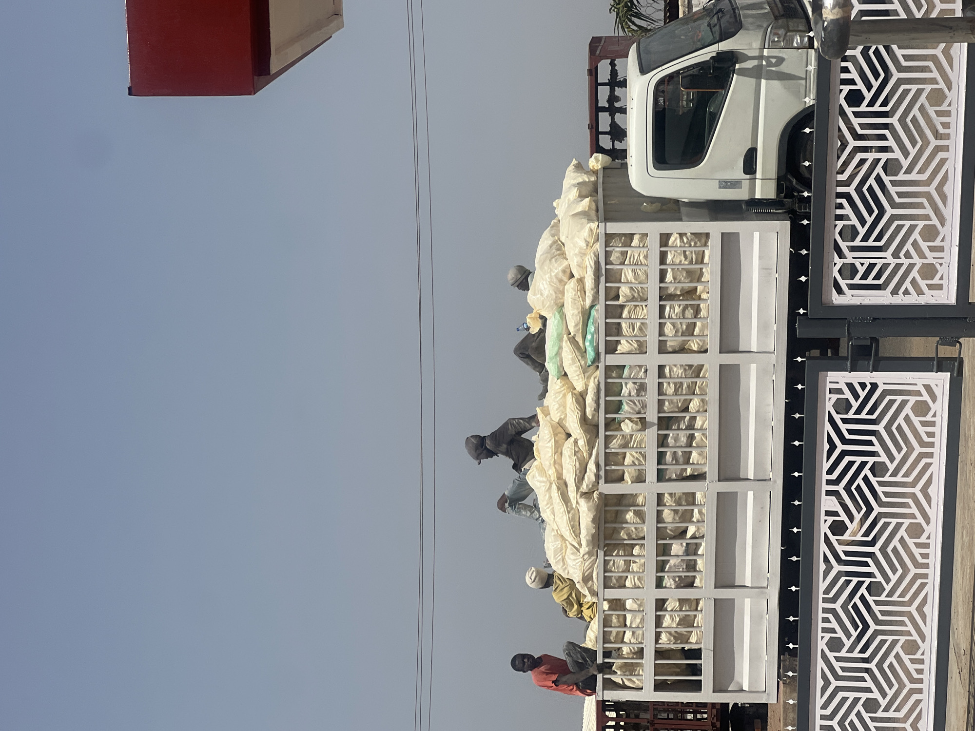 A medium shot depicts a light-duty truck loaded with sacks, with several people sitting on top of the load. The truck is parked next to a geometric patterned gate. The setting is likely urban in Barakin Lerawi, Nigeria, as suggested by the presence of the truck and the gate. The people on the truck are likely laborers involved in transporting the sacks. The sacks appear to contain an unknown material, potentially agricultural goods. The scene suggests manual labor and goods transportation.
