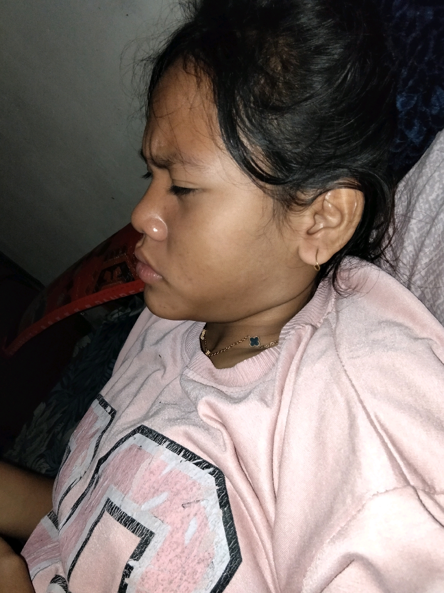 A close-up, profile shot of a young person's face and upper body is shown. The person is lying down, facing left, with their eyes closed. They have dark hair pulled back, and are wearing a pink t-shirt with a graphic design. A gold chain with a small dark blue pendant is visible around their neck. The background is dark and appears to be indoors, possibly a bedroom. The image was taken in Perambahan, Indonesia.