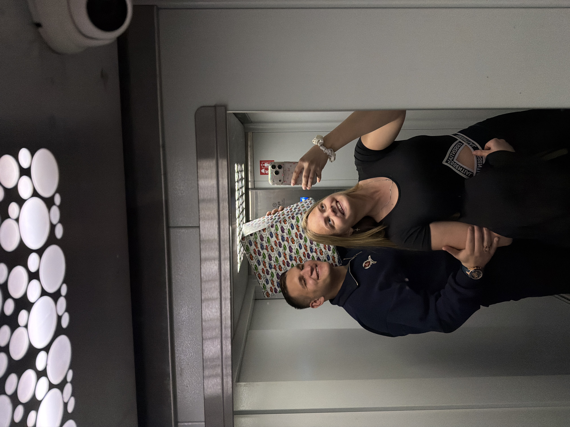 A couple is taking a selfie in what appears to be an elevator. The woman has blonde hair and is wearing a black top. She is holding a phone to take the picture. The man is wearing a dark blue jacket with a small emblem on the chest. They are holding a gift wrapped in paper with a colorful pattern. The elevator has a metallic interior. The background shows an artistic panel with circular cutouts and a security camera mounted on the wall. The location, based on the provided information, is Izhevsk, Russia.