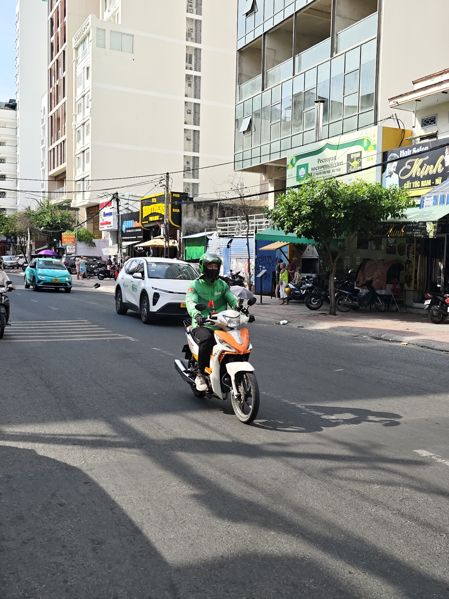 The image depicts a street scene in Nha Trang Ward, Vietnam, on a bright, sunny day. A person wearing a green helmet and a green jacket, presumably a delivery driver for "Grab," is riding a white and orange motorcycle. The driver is looking forward, and the motorcycle is in motion. In the background, a white electric car with "VinFast" branding is visible, also in motion. A turquoise electric car is parked to the left of the road. The street is lined with buildings, including a modern glass-fronted building and other multi-story structures. Several shops are visible on the street level, with signage in Vietnamese, including a "Hair Salon." The road shows clear lane markings and a pedestrian crossing. The overall environment is that of a busy urban street with vehicle traffic and commercial establishments. Shadows cast by trees and vehicles are visible on the road surface, indicating the direction of the sun.