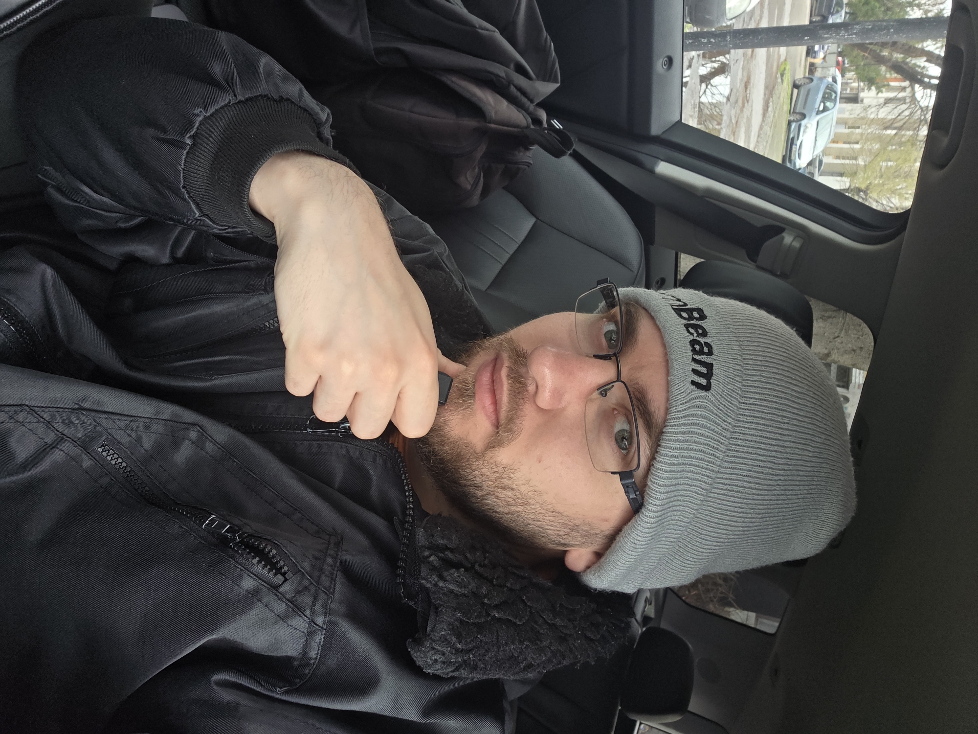 A man is sitting in the driver's seat of a car. He's wearing a black jacket with a fuzzy collar and a grey beanie with "Beam" printed on it. He has glasses on. He is holding a small, black, rectangular object between his fingers. The car is parked, and through the window, it's possible to see buildings and cars outside. The location cannot be determined to be in Plovdiv, Bulgaria from this image.