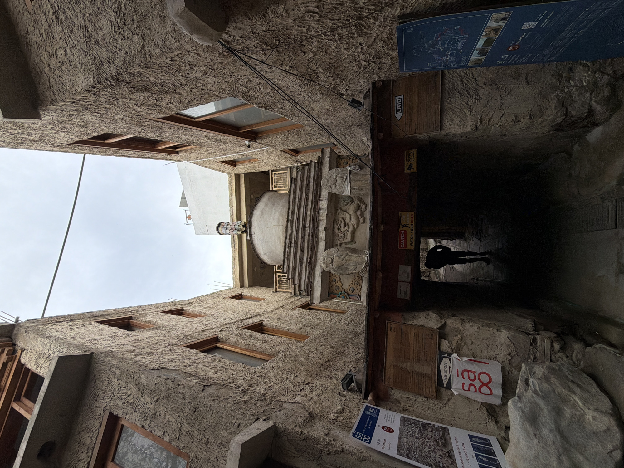 This is a low-angle, wide shot of a narrow alleyway in Leh, India, leading towards a Buddhist stupa. The walls of the alley are made of rough, textured stone with wooden window frames and balconies. A person is seen walking away from the camera into the darker section of the alley. Above the alley entrance, there is a decorated section with carvings and a stupa. Signs are visible on the walls, including a poster in English and a sign with "sa ∞ l" logo, possibly indicating a shop or studio. The sky is overcast.