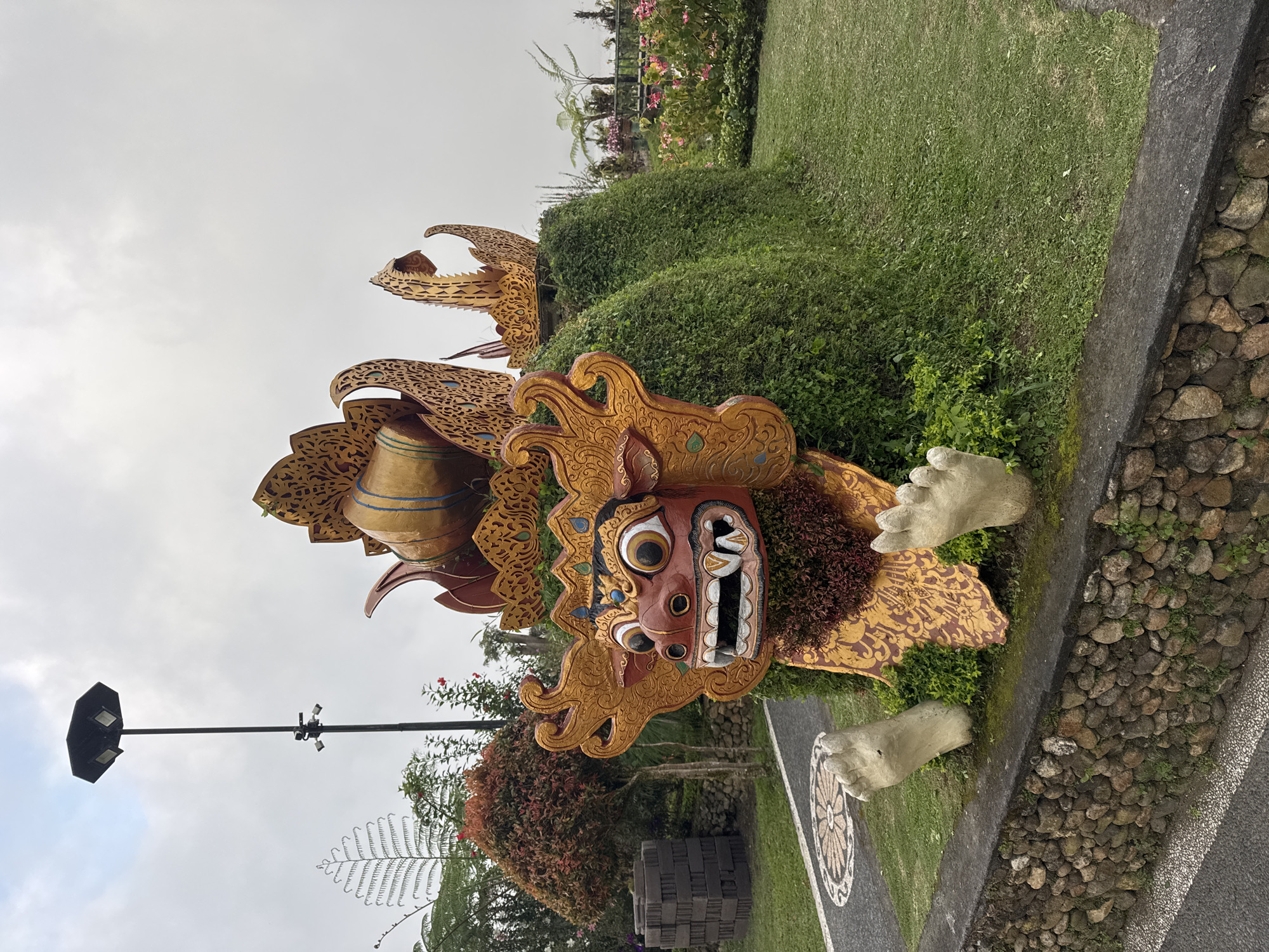 The image shows a Barong statue in a park or garden setting. The statue has an ornate mask with a gaping mouth, prominent eyes, and decorative patterns. It is gold and red, with green foliage around it. In the background is a grassy area with other plants and flowers, as well as a stone pathway. The sky is overcast. A lamppost is also visible in the background. The location is unknown.