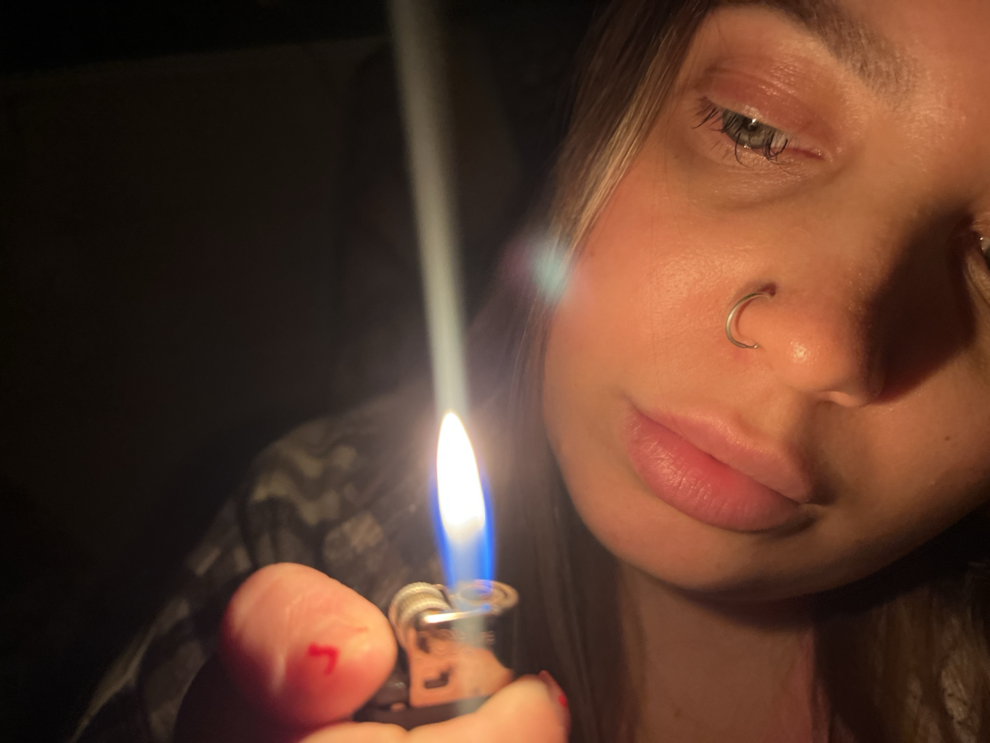 A close-up shot captures a person holding a lit lighter. The flame is bright blue and white, illuminating the person's face. The person has a nose ring and appears to have painted red marks on their fingernail. The background is dark, with hints of patterned fabric visible. The overall lighting suggests a low-light environment, possibly indoors at night. No specific location in Novopokrovskaya, Russia, can be determined from the image.