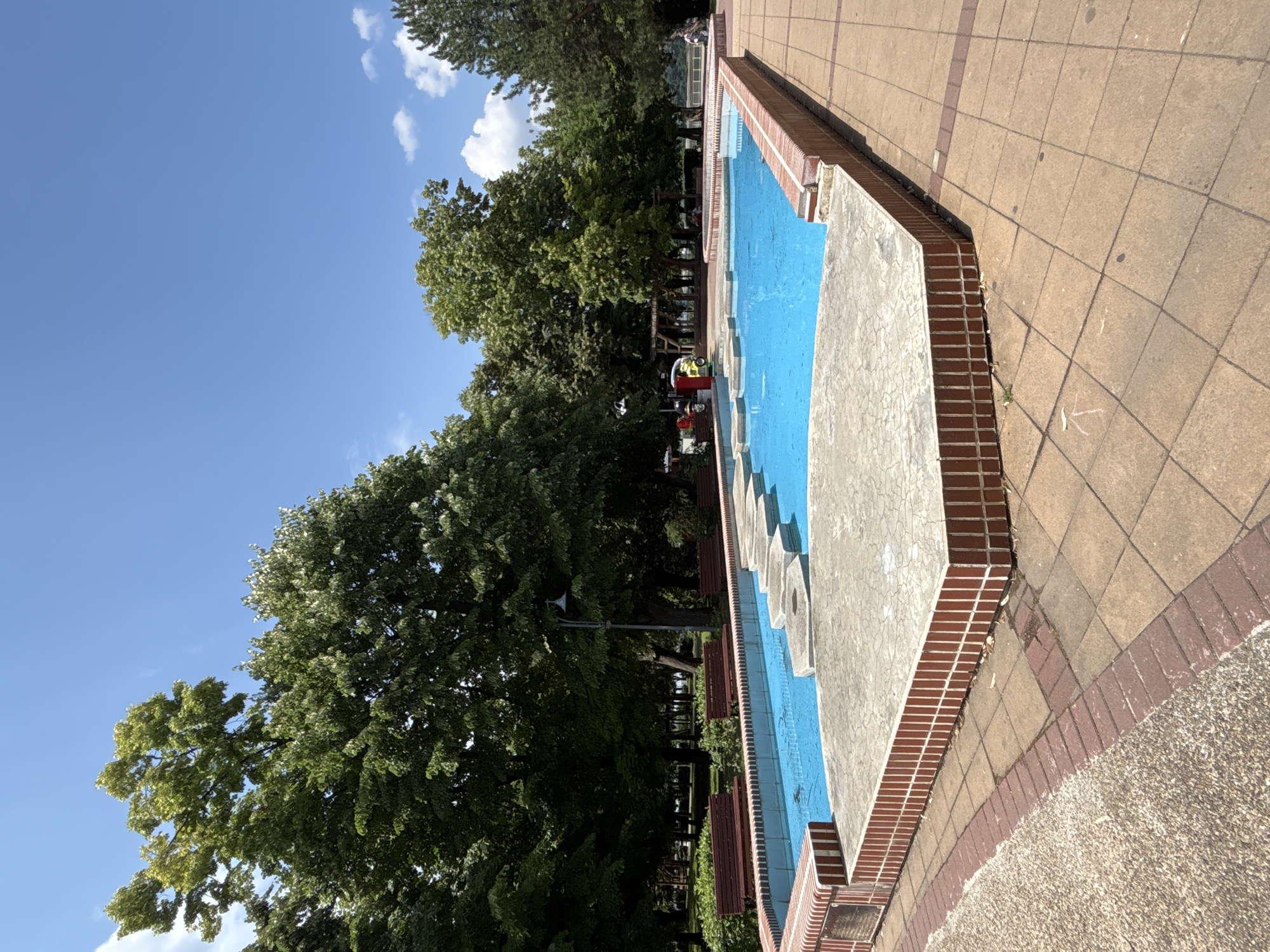The image depicts a public park scene in the city of Silistra, Bulgaria, featuring a shallow, rectangular swimming pool with bright blue water. The pool is surrounded by a tiled walkway and a brick border. Within the pool, there are several flat, grey stepping stones that create a path across the water. To the left of the pool, a row of red park benches is visible, and beyond them, a line of mature trees with lush green foliage. A lamppost stands near the benches. In the background, more trees and some park structures can be seen, with a glimpse of a body of water in the distance. The sky is clear and blue with a few scattered white clouds. In the far right of the image, a person is seated on a bench.
