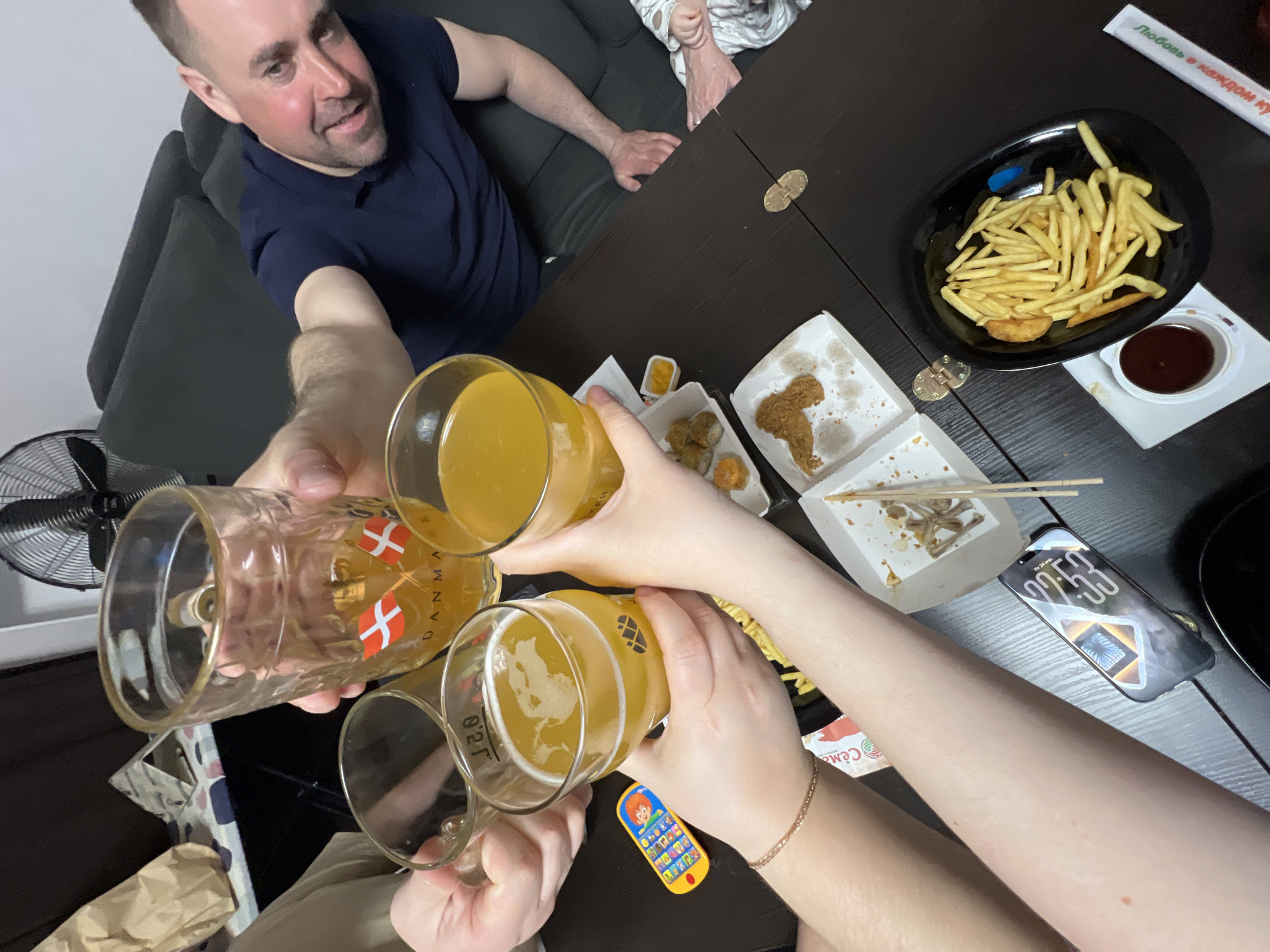 A high-angle shot depicts a convivial gathering around a dark-colored table, likely in an apartment in Perm, Russia. Four individuals are toasting with beer glasses, two of which feature Danish flag decals, filled with a yellow liquid. The visible participants include a man in a navy blue polo shirt in the upper left, and the hands of three others are engaged in the toast. The table setting includes a black dish filled with french fries, condiment containers, open food containers with leftover chicken pieces, sushi, and wooden chopsticks. A smartphone is placed on the table, partially obscuring the surface, with the screen visible. A small, colorful toy phone is also present. A fan can be seen in the background to the left, and parts of a couch and other domestic items are visible, giving context to the indoor setting. The location is identified as Perm based on the Cyrillic inscription seen on a condiment container.