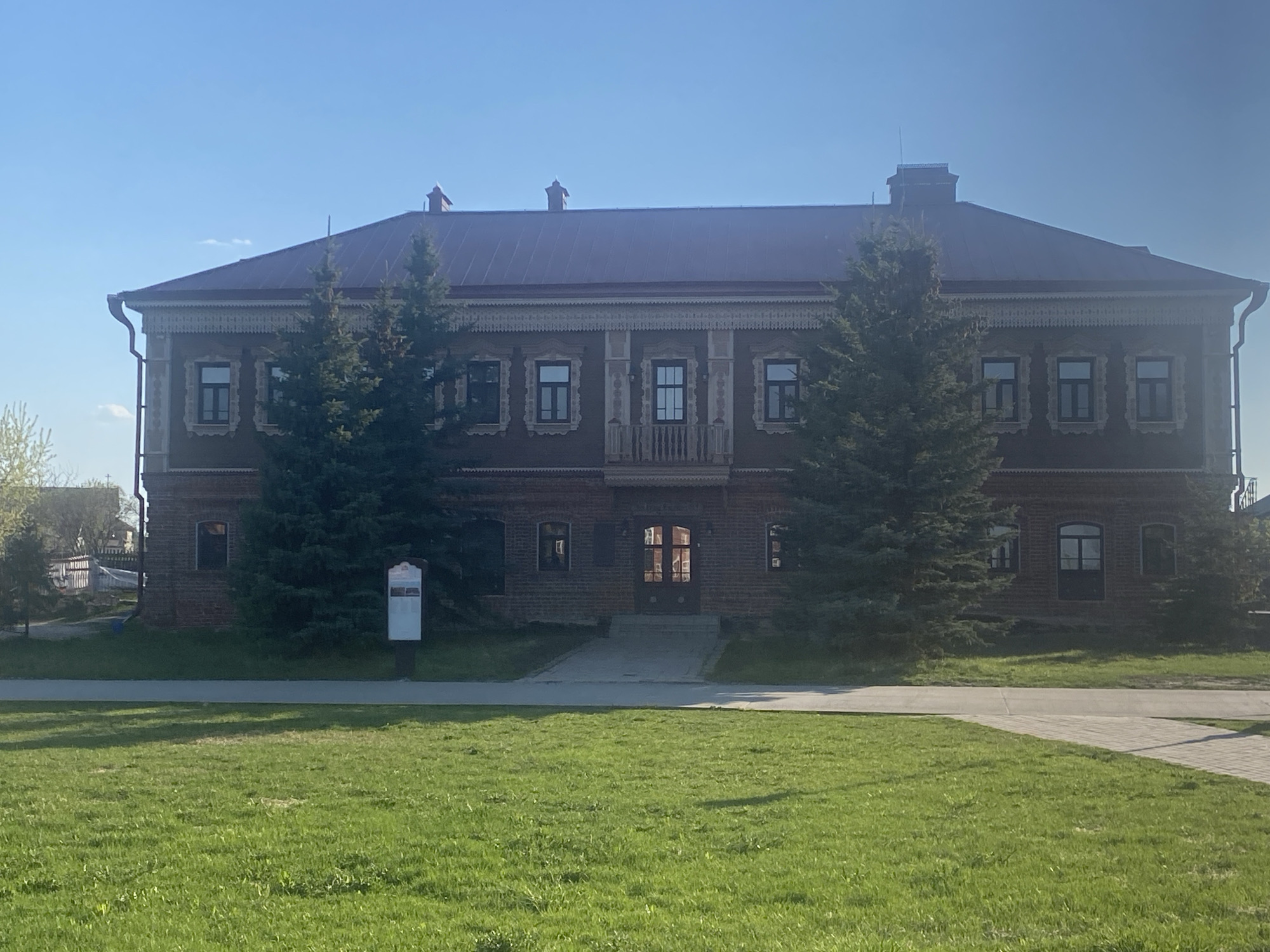 The image shows the building of the Sviyazhsk History Museum, located in the city of Sviyazhsk, Russia. The two-story building is made of red brick and has a dark brown roof. Two tall evergreen trees partially obscure the building's facade. A sign is visible to the left of the entrance path. A green lawn stretches in front of the building. The sky is clear and blue.