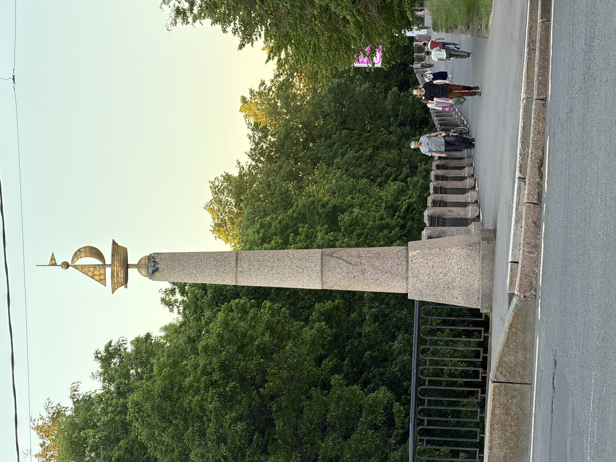 This image depicts the Lomonosov Bridge over the Fontanka River in Saint Petersburg, Russia, characterized by one of its distinctive granite pylons topped with a gilded sphere holding a model of a sailing ship and a small flag. The scene is captured on a paved bridge with a gray asphalt roadway featuring a faded yellow dividing line in the foreground. To the right, a raised concrete sidewalk with a rough-hewn, light reddish-brown granite curb separates the pedestrian area from the road. The bridge features a dark, ornate metal railing with regularly spaced balusters and arched top sections, which extends along the sidewalk. A prominent, tapered granite column, constructed from stacked blocks of mottled pinkish-gray granite, stands on the right side of the sidewalk. Its base is wider and roughly trapezoidal. The column culminates in a dark spherical base, supporting a large, ornate golden spherical finial, which in turn holds a stylized golden model of a sailboat with a single mast and a small triangular flag pointing right. Evidence of dark staining is visible on the column directly beneath the golden sphere. Several pedestrians are visible walking on the sidewalk. In the mid-ground, a man with short grey hair, wearing a light grey t-shirt and dark trousers, walks towards the viewer, pushing a dark-framed bicycle with a light seat. Slightly to his right and closer to the viewer, another man with long dark hair, wearing a dark patterned t-shirt and brown trousers, walks directly towards the viewer, carrying a green object in his left hand. Further back on the sidewalk, a woman in a light-colored top and what appears to be a pink skirt or trousers walks away from the viewer. Behind her, two men also walk away: one in a white t-shirt and light trousers, and another in a dark t-shirt and light trousers. A fifth, indistinct figure is visible further down the bridge. Lush green deciduous trees with full canopies line both sides of the bridge, creating a verdant backdrop. The tops of some trees show bright yellow-green highlights from the sunlight. In the distance on the right, partially obscured by trees, a bright magenta-pink light source, possibly a digital display or sign, is visible. The sky above is a clear gradient from light blue at the top to a pale yellow-green closer to the horizon, indicating either early morning or late afternoon light. A thin, dark power line