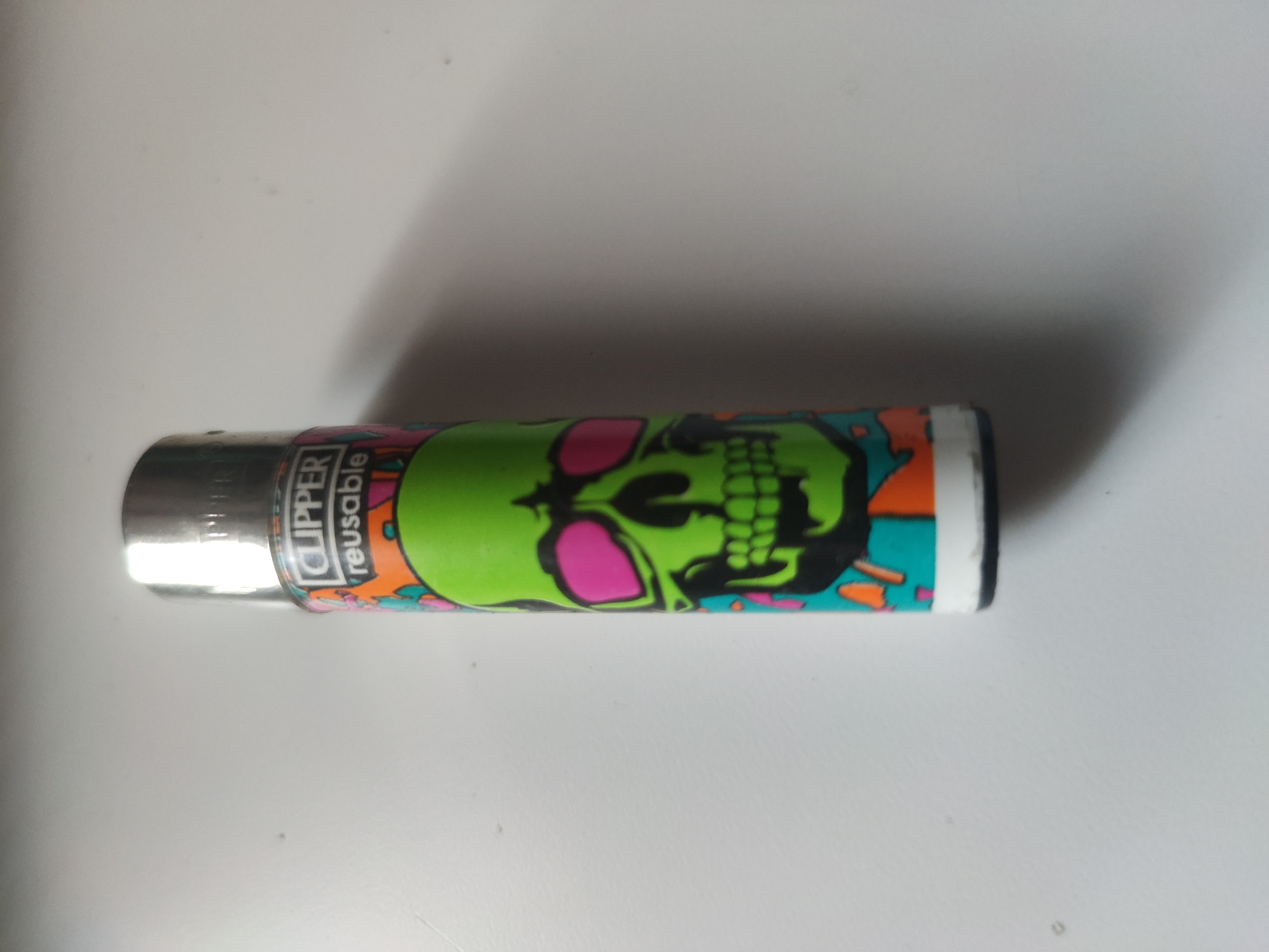 A close-up shows a Clipper brand lighter with a skull design lying on a white surface. The skull is predominantly green with pink eye sockets and black shading. The lighter's body is also decorated with other colorful patterns. The metallic top of the lighter bears the Clipper logo and the word "reusable."