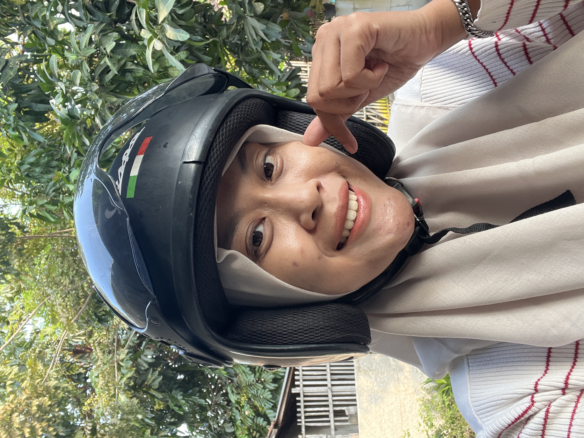 A woman wearing a white hijab and a white sweater with red stripes is taking a selfie. She is also wearing a black helmet with Italian flag colors on it. She is smiling and pointing to her cheek with her right hand. The background of the image is out of focus and shows trees and a fence. The scene appears to be outdoors, possibly in a residential area.