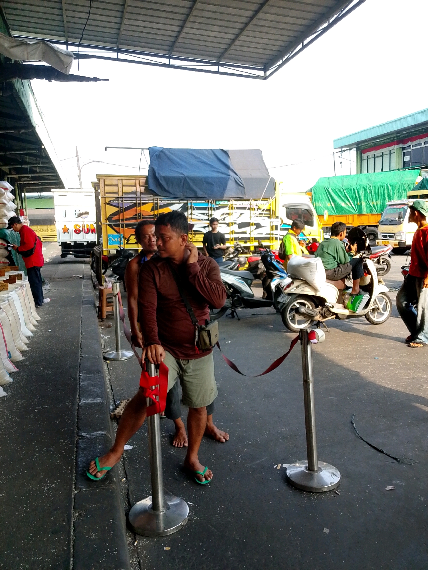 The image depicts an outdoor scene, likely a market or loading area, in East Jakarta, Indonesia. In the foreground, a man wearing a maroon long-sleeved shirt and khaki shorts stands with his hand resting on a red barrier tape attached to a stanchion. To his left, another man in a dark shirt and patterned shorts stands behind him, partially obscured. Behind them, several large trucks are parked, some covered with tarps. One truck has colorful artwork on its side. In the background, a person in a red shirt is interacting with stacked white sacks, possibly rice, along a covered walkway. Several motorcycles are parked in the area, and people are milling around. The ground is paved and appears to be a public thoroughfare. The lighting suggests it is daytime, with sunlight casting shadows.
