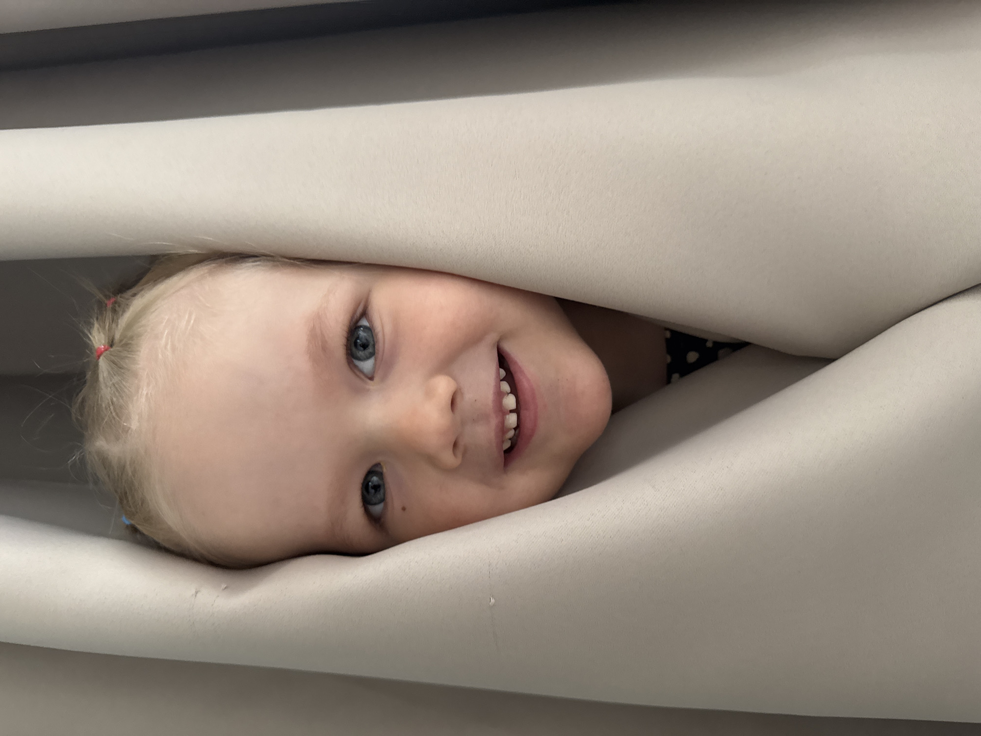 A close-up, horizontal image features a young child peeking out playfully from between light-colored, soft material folds. The child, positioned centrally and looking upwards towards the viewer, has fair skin, blonde hair, and striking blue eyes. A small red hair tie is visible on the left side of their head. They are smiling broadly, revealing their teeth, and their expression conveys joy and amusement, suggesting a game of peek-a-boo or hiding. The material surrounding the child appears to be fabric, possibly upholstery or bedding, with a fine woven texture. It forms a curved, enveloping space around the child's head. The lighting is bright and even, indicating an indoor setting. No text is visible in the image.