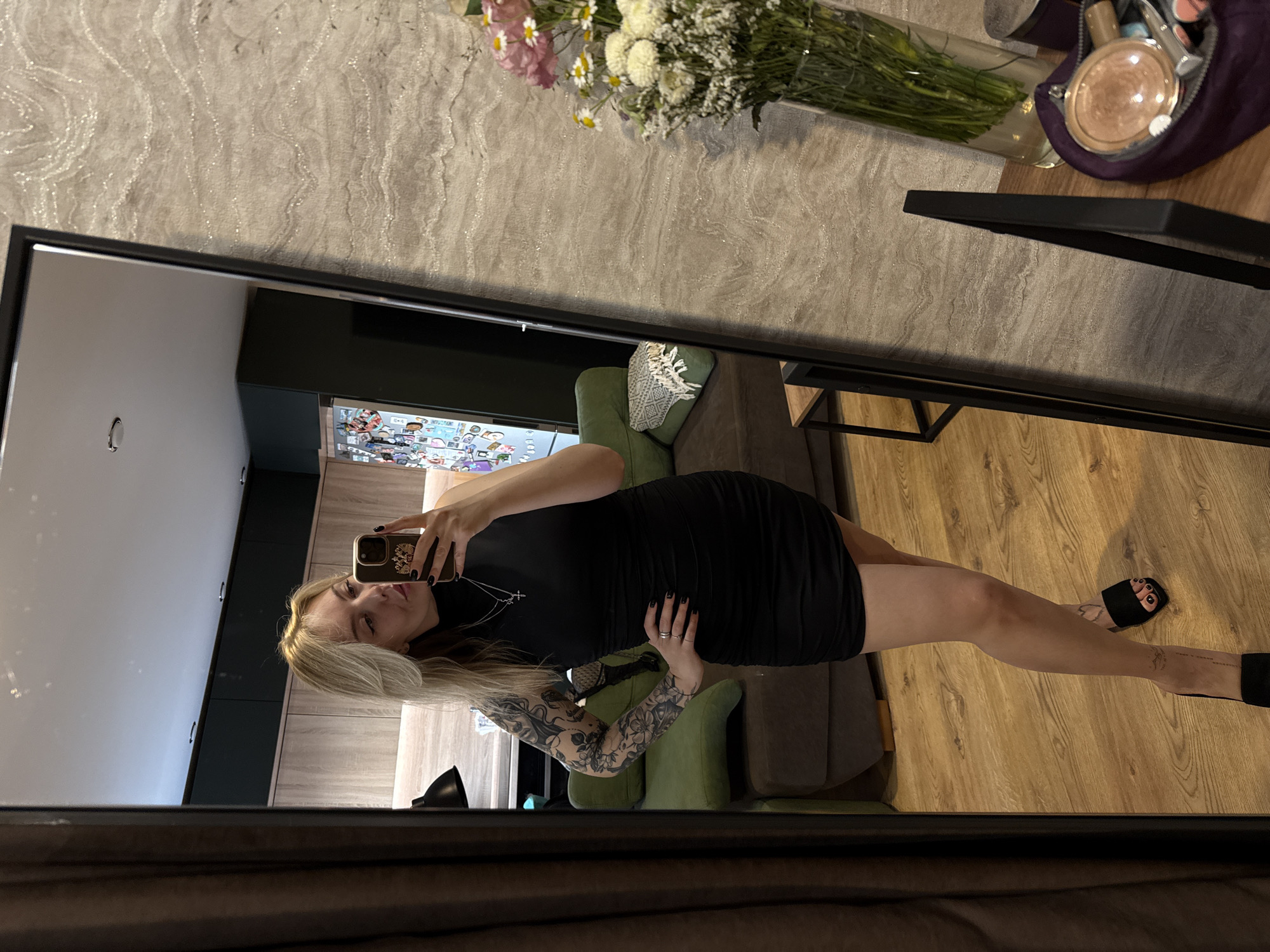 A woman stands in front of a full-length mirror, taking a selfie. She is wearing a black dress and black sandals. She has blonde hair and tattoos on her arm. The reflection shows a living room with a green couch, a vase of flowers, and a vanity with makeup. In the background is the reflection of the kitchen with visible stickers on the fridge. The photo appears to be taken indoors. There are no indications that this image was taken in the city of Krasnoyarsk, Russia.