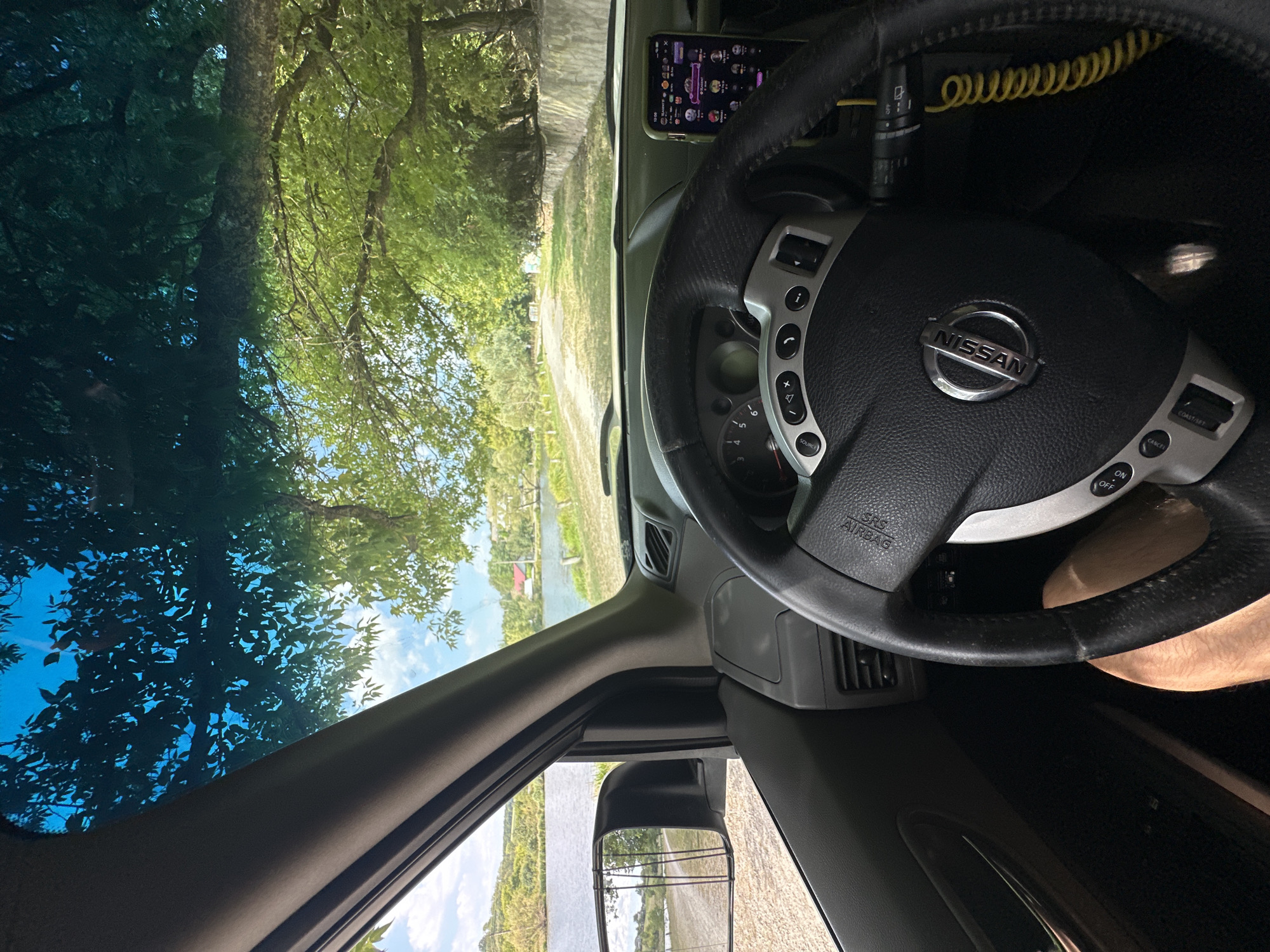 The image is taken from the interior of a car, looking forward through the windshield. The driver's hands are not visible, but a leg is partially visible, indicating a person is in the driver's seat. The steering wheel, dashboard, and a smartphone mounted on the right side of the dashboard are prominent. The view through the windshield shows a dirt road leading towards a body of water, likely a lake or river, with trees and buildings on the far side. The sky is blue with some clouds. The scene is set in a natural, rural environment. There are no discernible features that would indicate the specific location within Makiv, Ukraine, as the image lacks any identifying landmarks or signage.